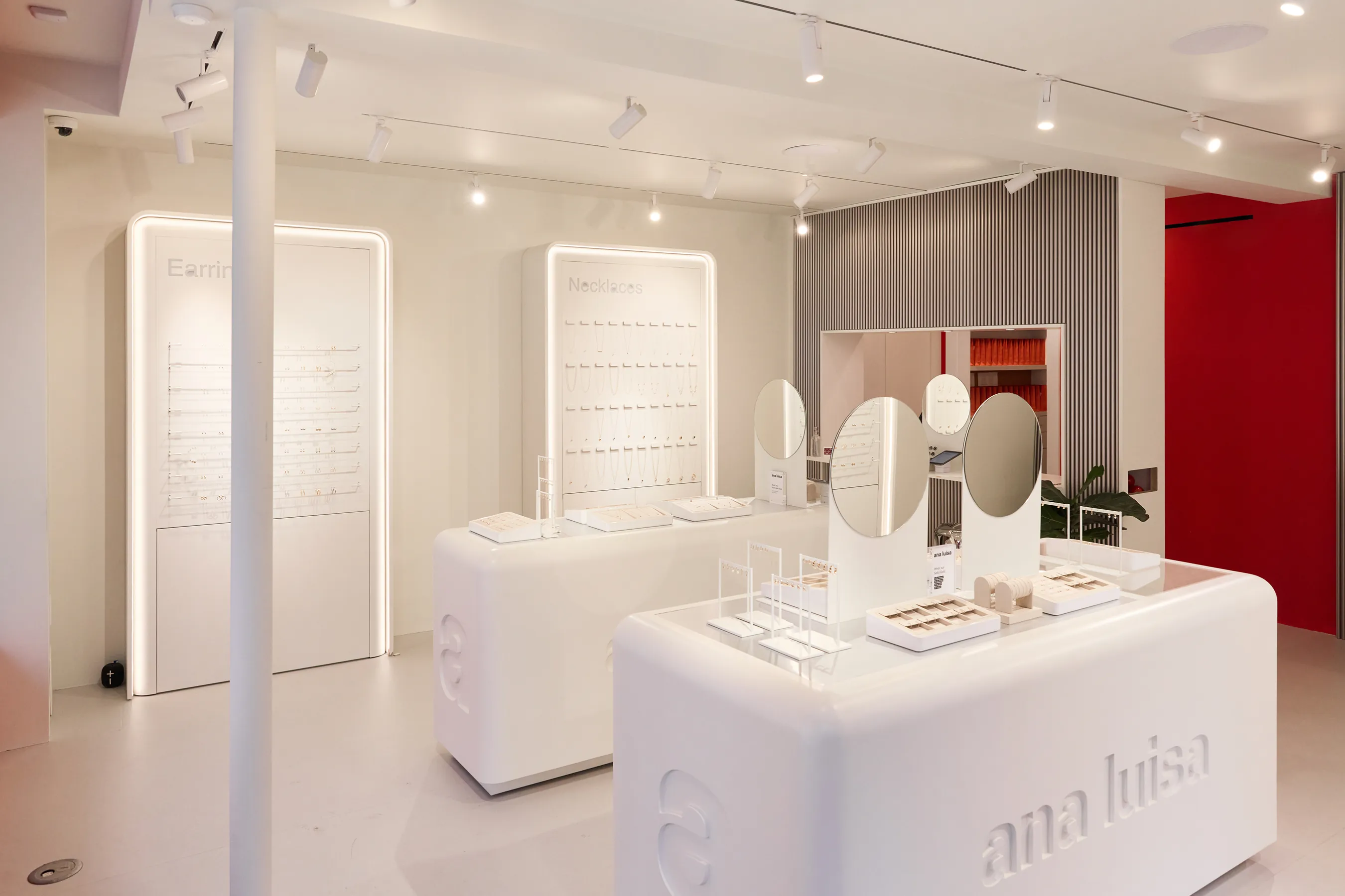 Interior of the Ana Luisa jewelry store featuring minimalist white display tables, circular mirrors, and illuminated wall panels showcasing earrings and necklaces. The clean, modern design highlights Ana Luisa’s sustainable, high-quality jewelry collection in a bright and inviting retail space.