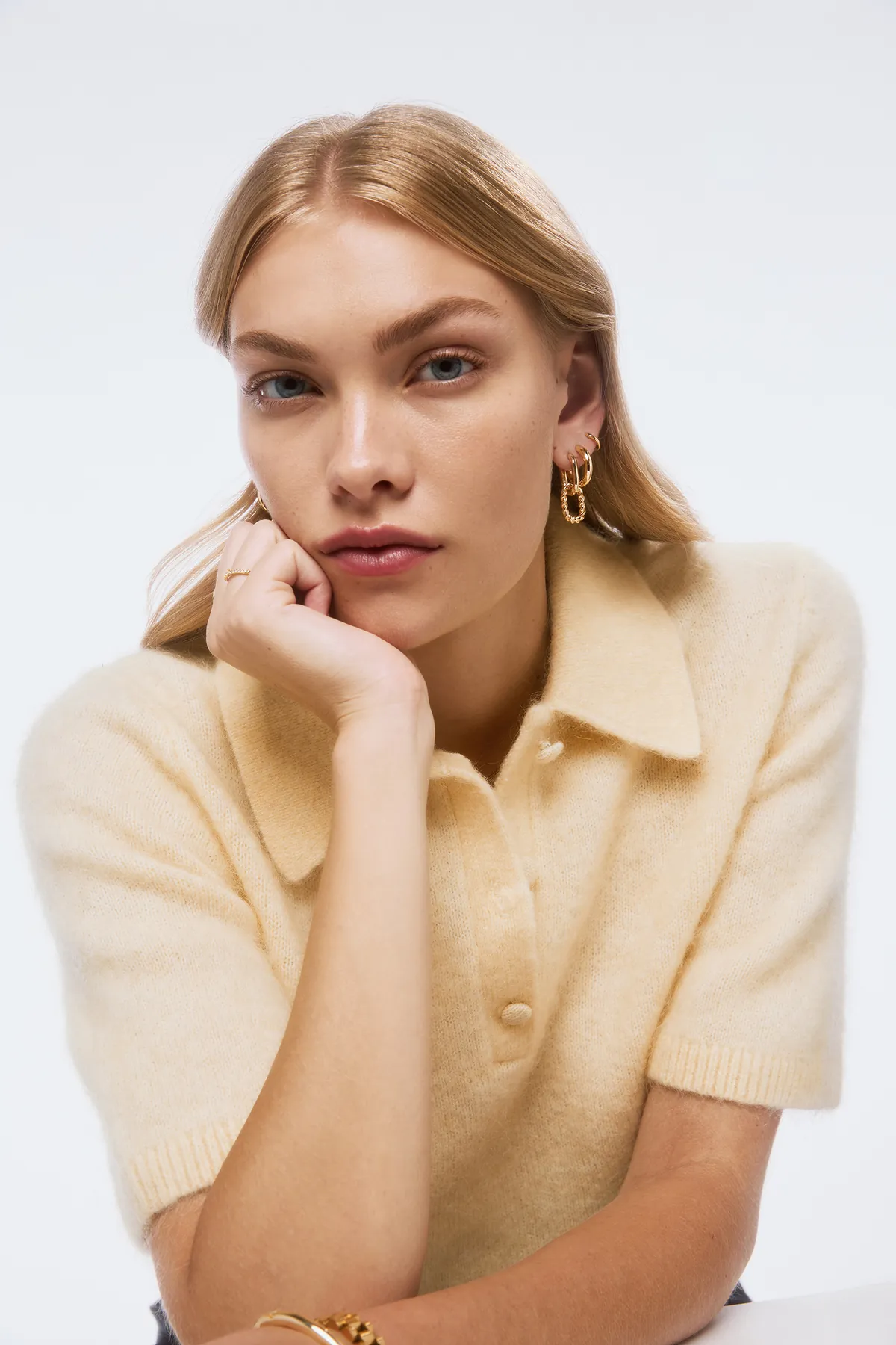 A model wearing gold hoop earrings, a gold ring, and a gold bracelet poses with her hand resting on her chin. She is dressed in a soft beige knit top with a collar, set against a clean studio background, highlighting Ana Luisa’s minimalist, elegant jewelry style.