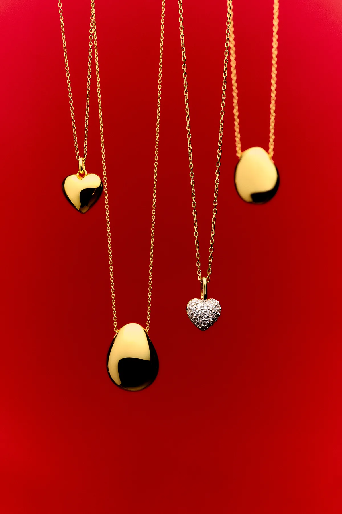 Gold heart and teardrop pendant necklaces from Ana Luisa displayed against a bold red background. The polished gold and pavé heart designs highlight the brand’s elegant, minimalist jewelry aesthetic.