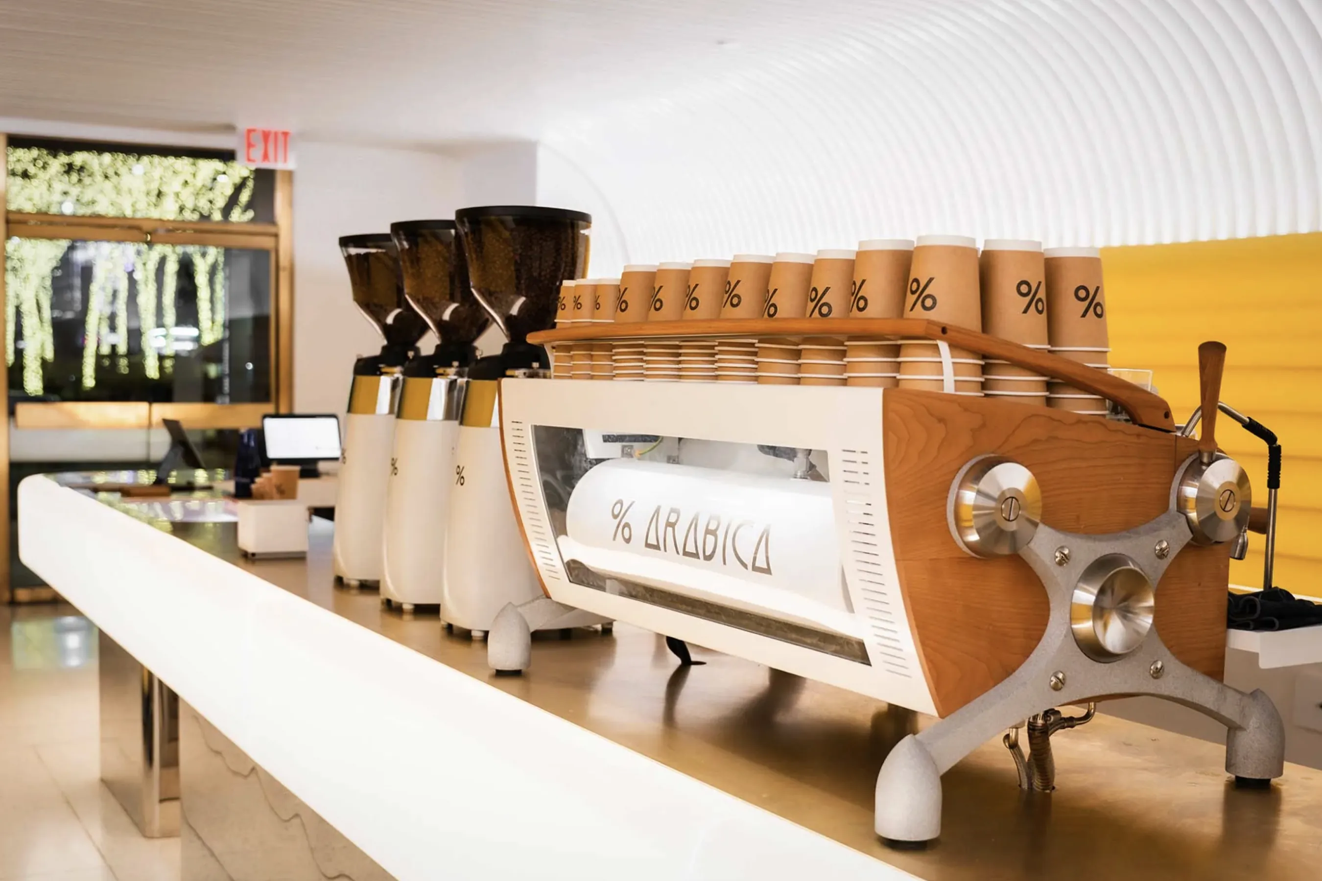 A modern coffee bar at % Arabica featuring a sleek white and wood espresso machine lined with branded brown cups displaying the % logo. Three large white grinders filled with coffee beans sit beside the machine on a minimalist counter. Warm lighting and a bright, contemporary interior frame the scene, with illuminated trees visible through the glass door in the background, highlighting % Arabica’s premium café aesthetic.