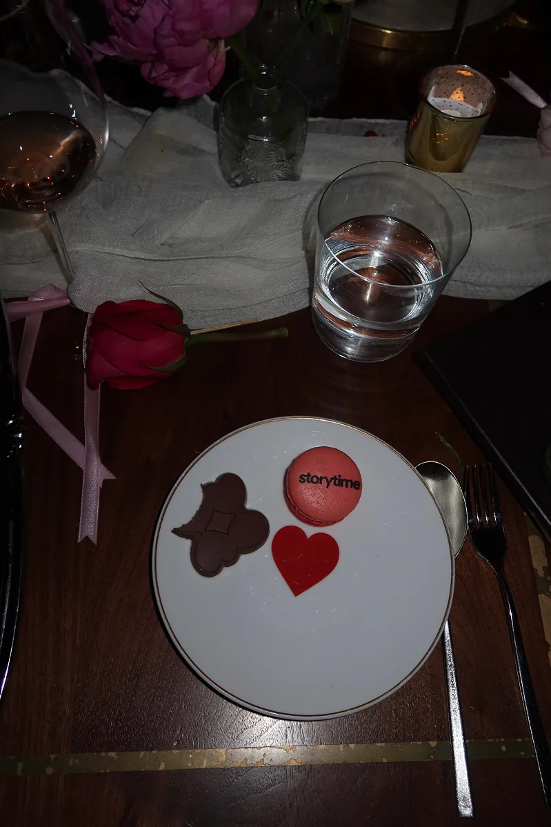 Storytime-branded macaron from @bklynmacs and Storytime branded heart-shaped Belgian chocolates served on a white plate at a candlelit Galentine’s dinner at Beefbar NYC, with pink roses and wine glasses on the table.