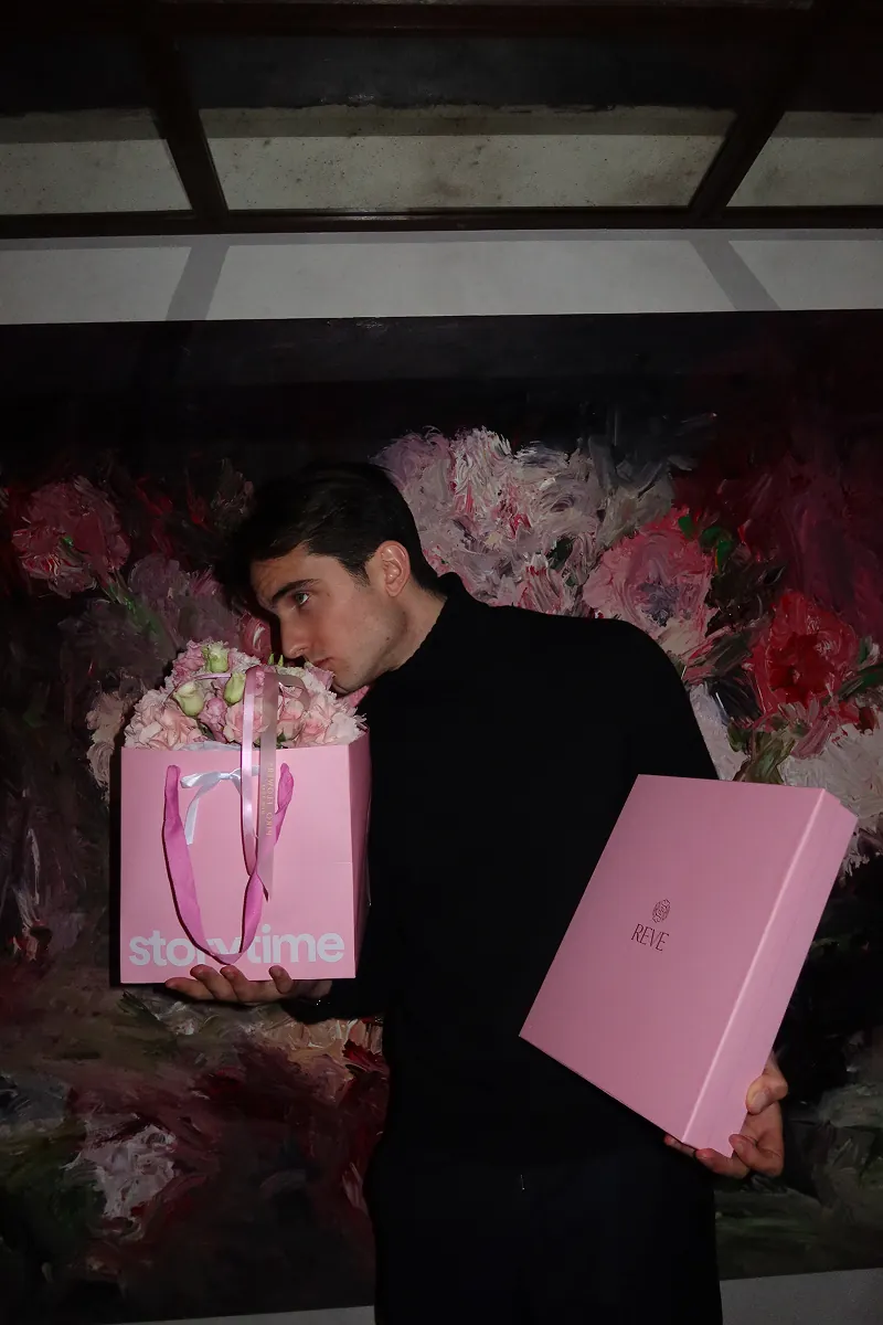@theeuropeankid dressed in black holding a pink Storytime floral gift bag from @piroflowers and matching gift boxes from @reve_sleepwear , posing in front of a wall of pink flowers at a Galentine’s dinner event in NYC.