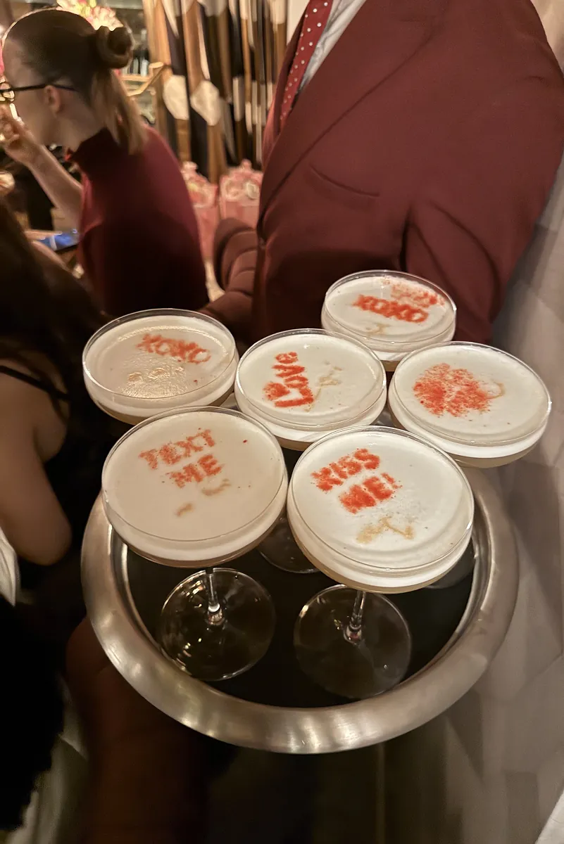 Server holding a silver tray of espresso martinis at Galentine’s Dinner at Beefbar NYC, each topped with red stenciled messages like “XOXO” and “KISS ME.”