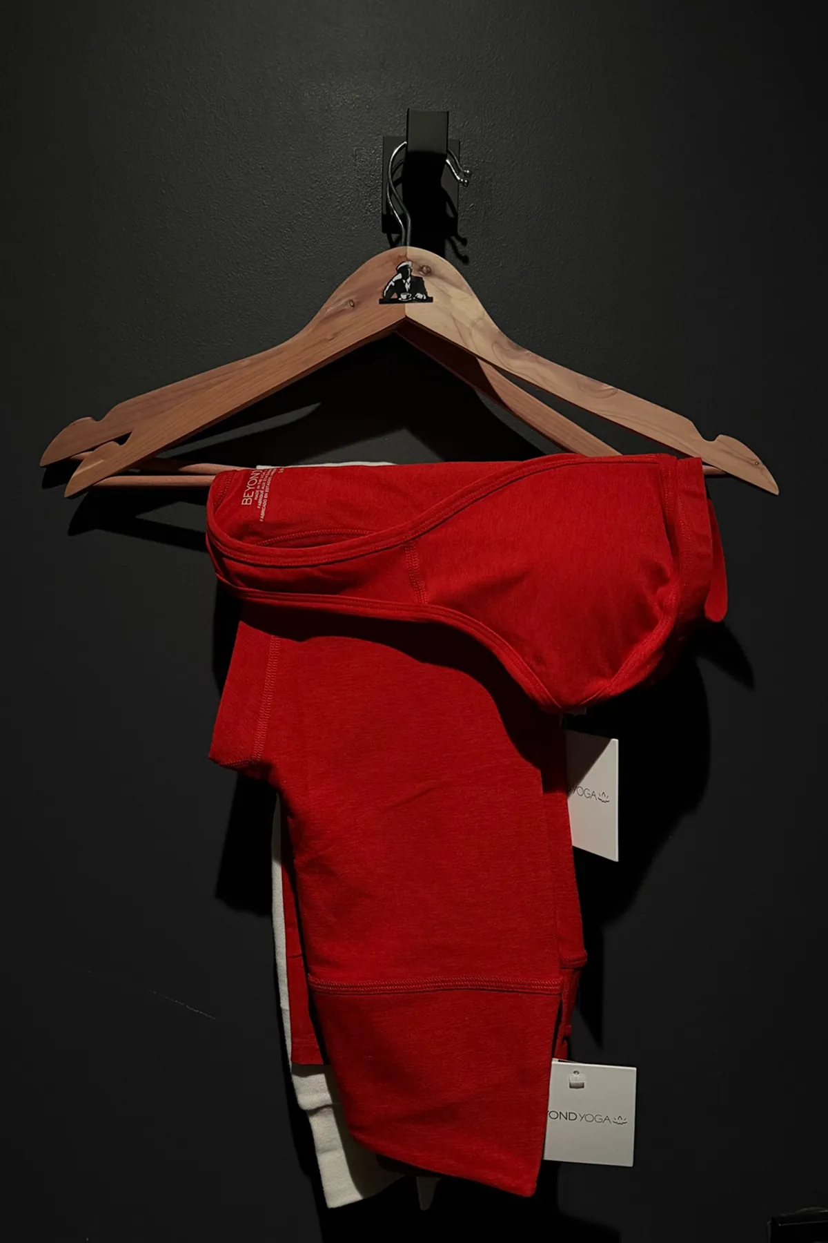 A red activewear set hanging on a wooden Dogpound-branded hanger against a dark wall, with tags visible from Beyond Yoga.