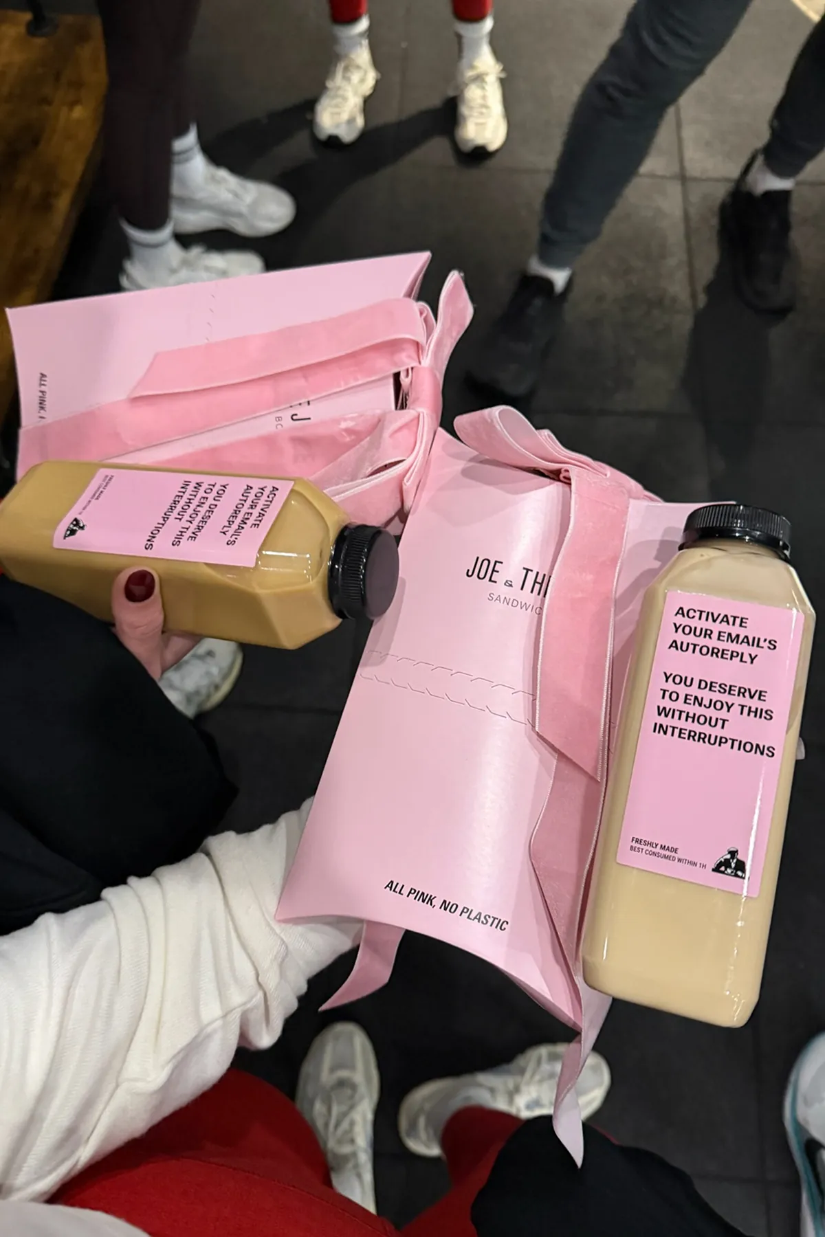Hands holding pink Joe & The Juice gift boxes tied with velvet bows and bottled juices with pink labels, with Storytime Creators in activewear and sneakers standing in the background on the gym floor.