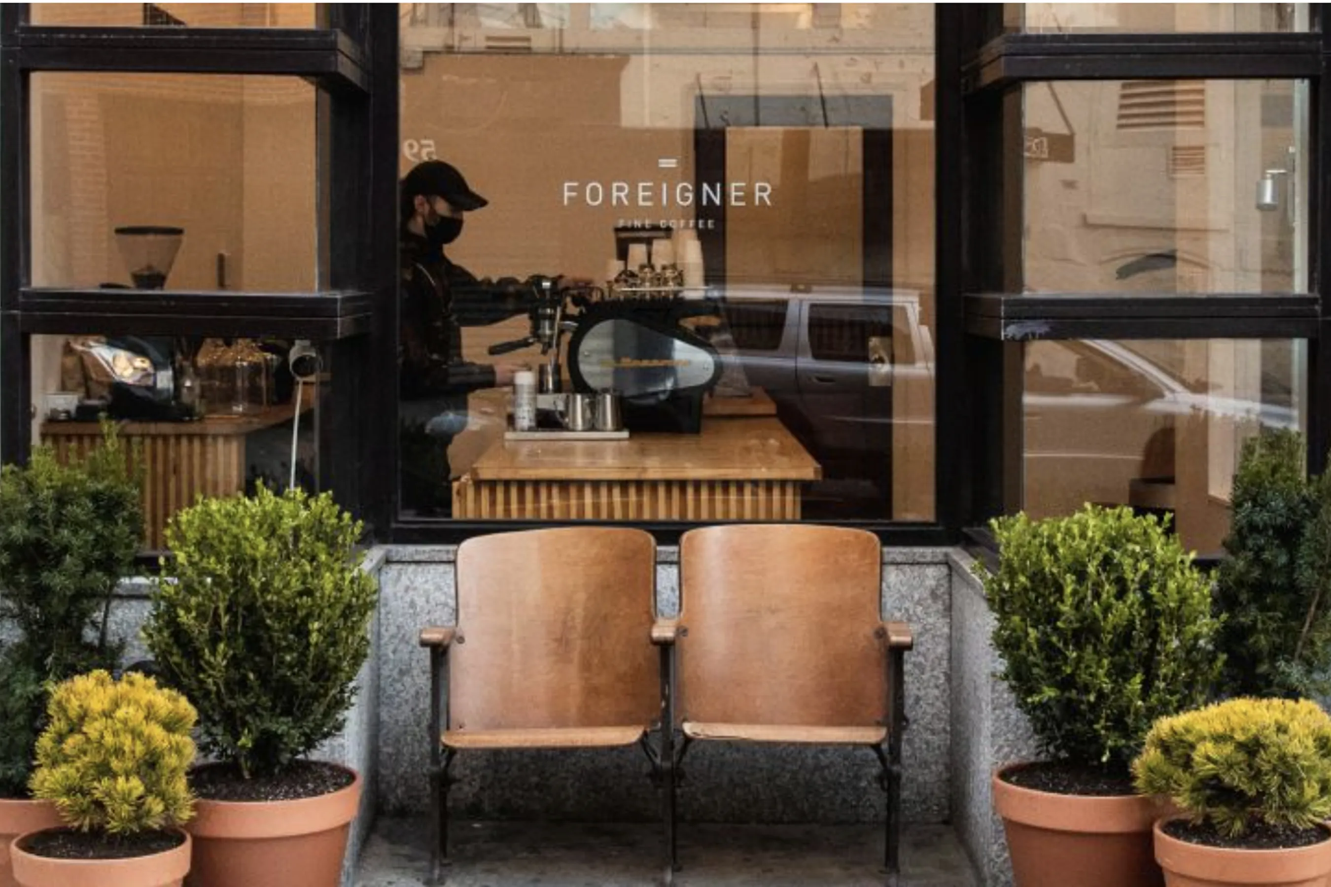 Storefront window of Foreigner Fine Coffee with a barista preparing drinks inside, visible through large glass panes. Two vintage wooden chairs sit outside surrounded by potted green plants, capturing the café’s minimalist, cozy, and design-forward aesthetic.