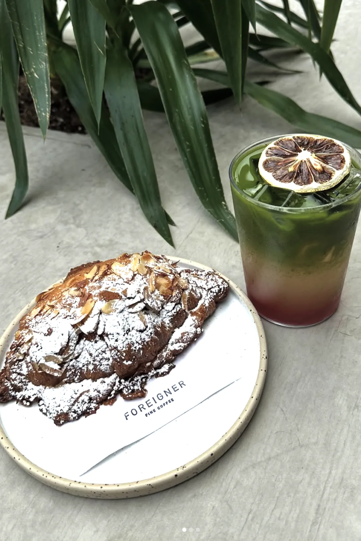 A powdered almond croissant served on a Foreigner Fine Coffee plate next to a layered iced matcha drink topped with a dried citrus slice. The setup sits on a light concrete surface with green plant leaves in the background, showcasing the café’s artisanal pastry and specialty beverage offerings.