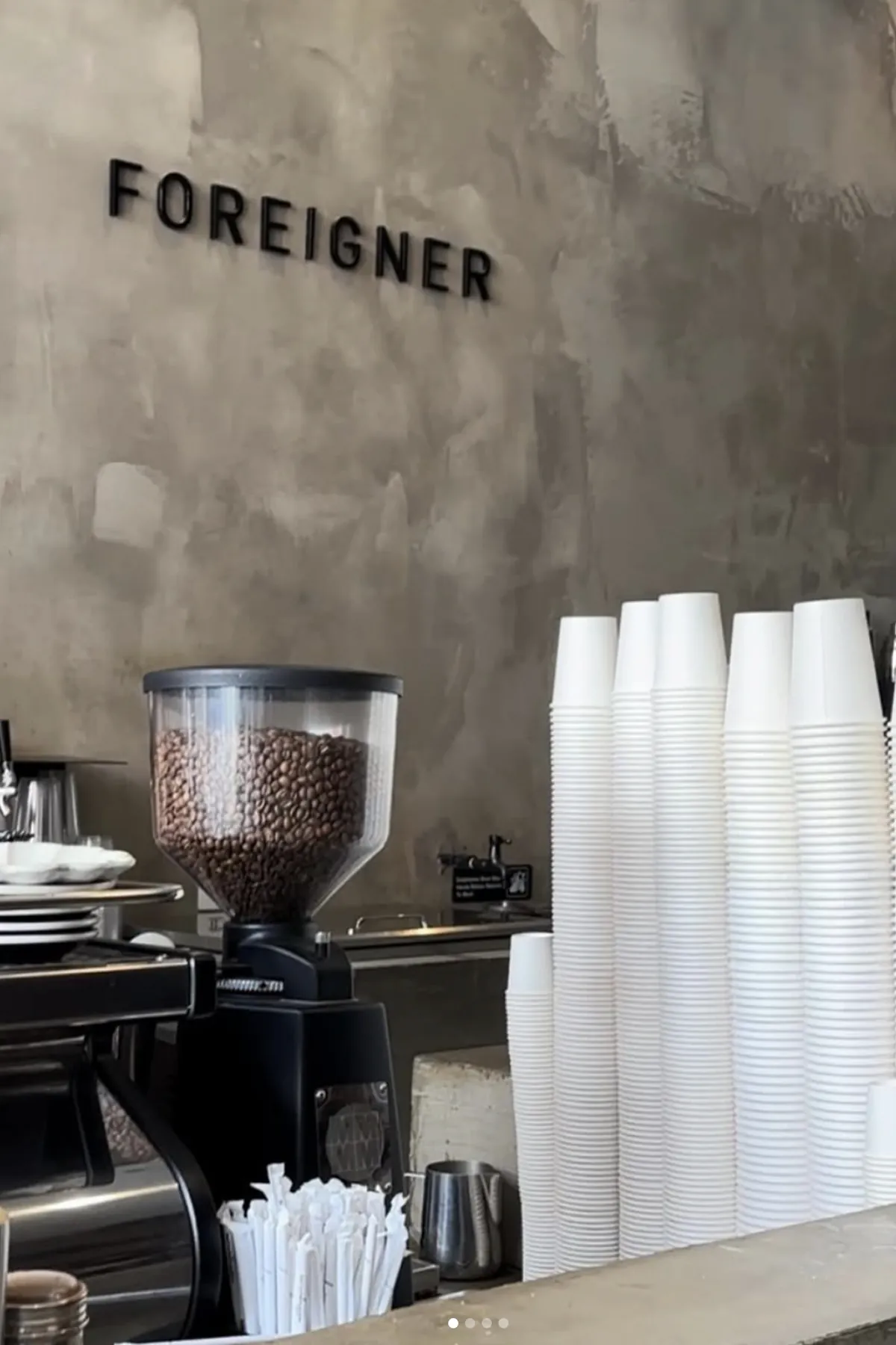 Interior of Foreigner Fine Coffee featuring a minimalist concrete wall with the “Foreigner” logo. A coffee grinder filled with beans sits beside neatly stacked white cups and barista tools, highlighting the café’s clean, modern aesthetic and specialty coffee focus.