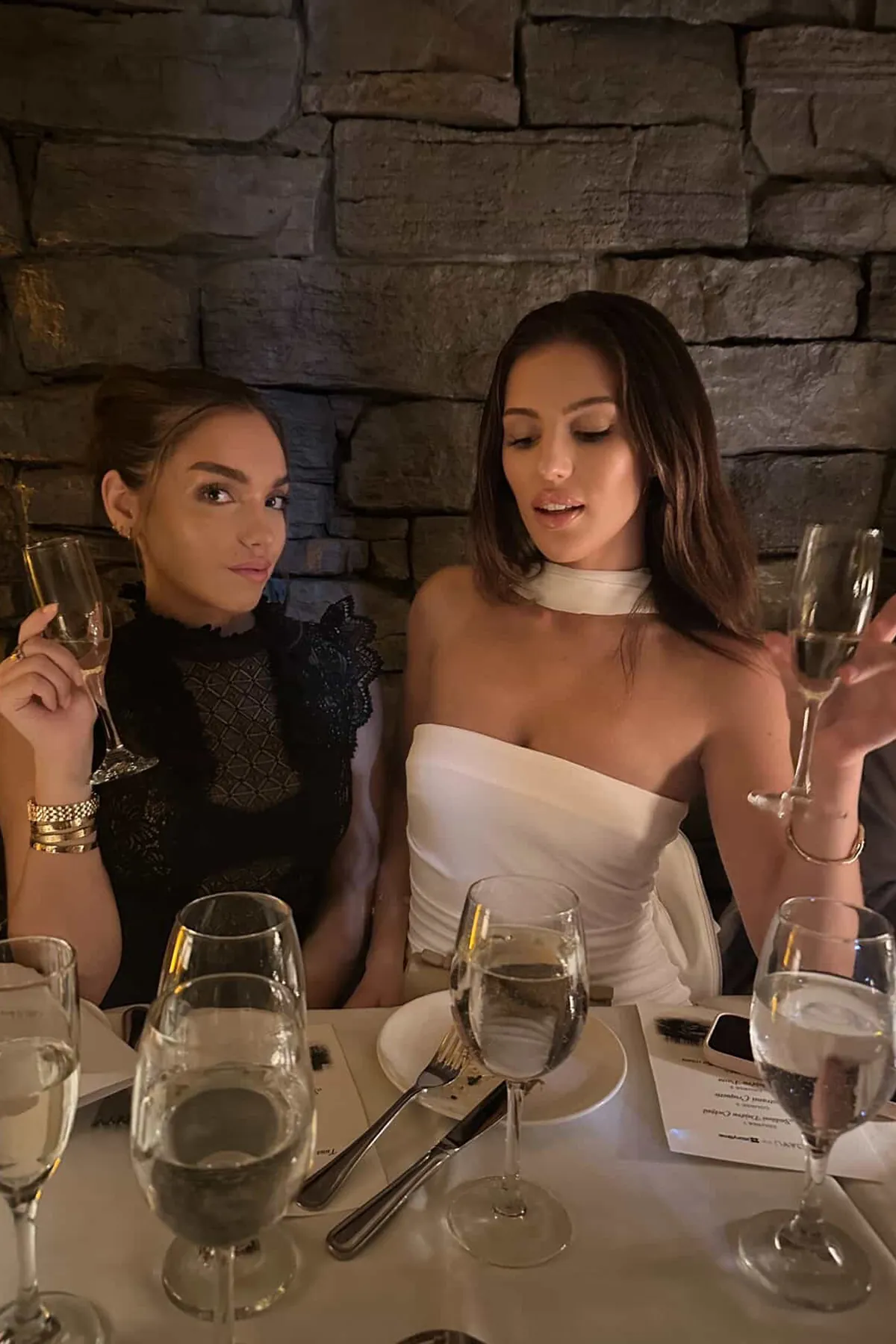 @anibollok and @chloeeleonikk seated at an intimate candlelit dinner table against a rustic stone wall, holding champagne flutes. One wears a black lace high-neck dress, the other a strapless white dress with a choker neckline. Multiple wine glasses and elegant place settings are arranged on the table, capturing a sophisticated, upscale dining atmosphere.