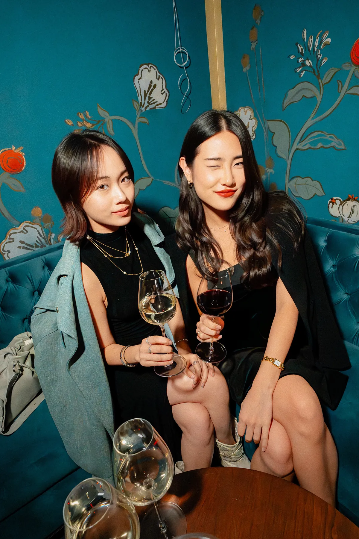 @_irenehwang sitting on a teal velvet couch at an event, holding glasses of wine and smiling, with a floral-patterned wall in the background and additional wine glasses on the table in front of them at Maison Barnes.