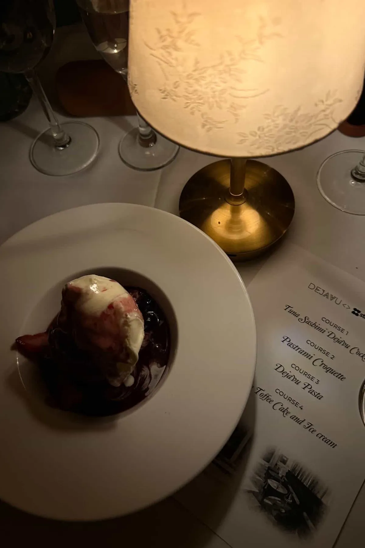 Close-up of a chocolate fondant dessert served in a white bowl under a softly glowing table lamp, with wine glasses and a printed “Déjà Vu x Storytime” multi-course menu beside it. The warm lighting and refined table setting highlight the intimate, elevated dining experience.