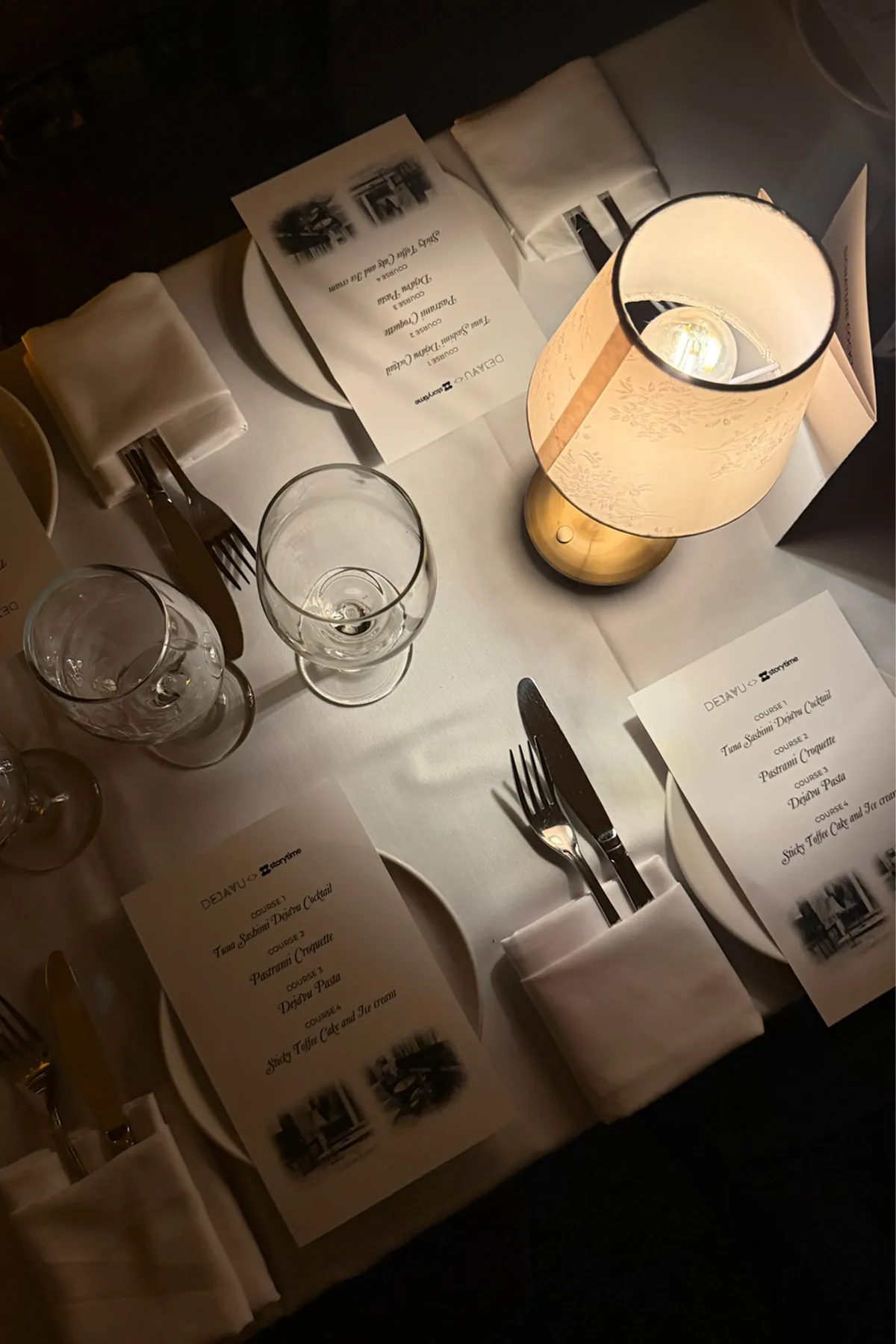 Overhead view of an elegantly set dinner table featuring white tablecloths, neatly folded napkins, polished cutlery, wine glasses, and printed multi-course menus branded “Déjà Vu x Storytime.” A softly glowing table lamp adds warm, intimate lighting, highlighting the refined and curated dining experience.