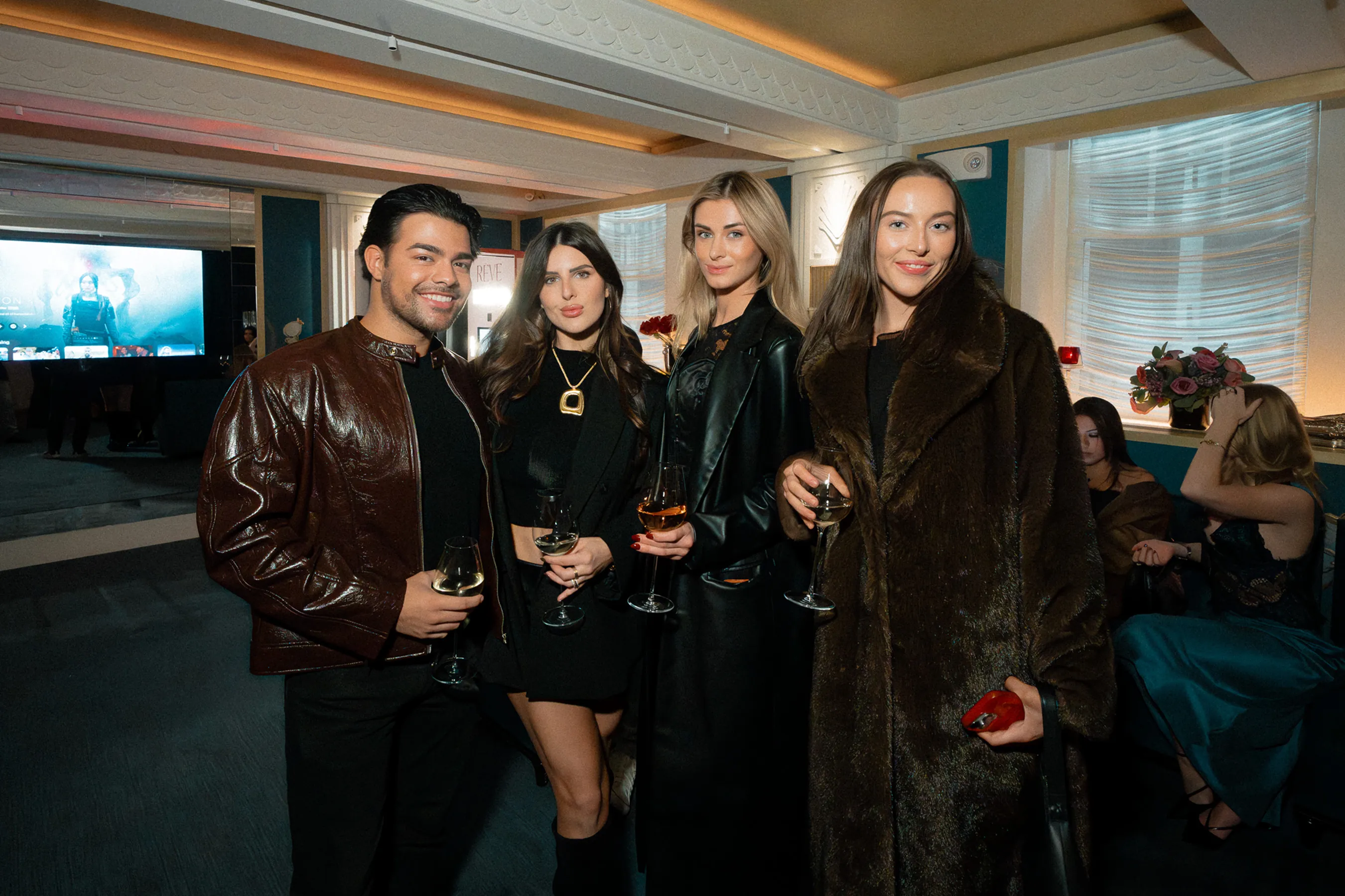 @becfic, @brian.ruballos, @_taniagrover, posing together at Maison Barnes, each holding a glass of wine. They stand in a warmly lit room with floral décor and additional attendees seated in the background.