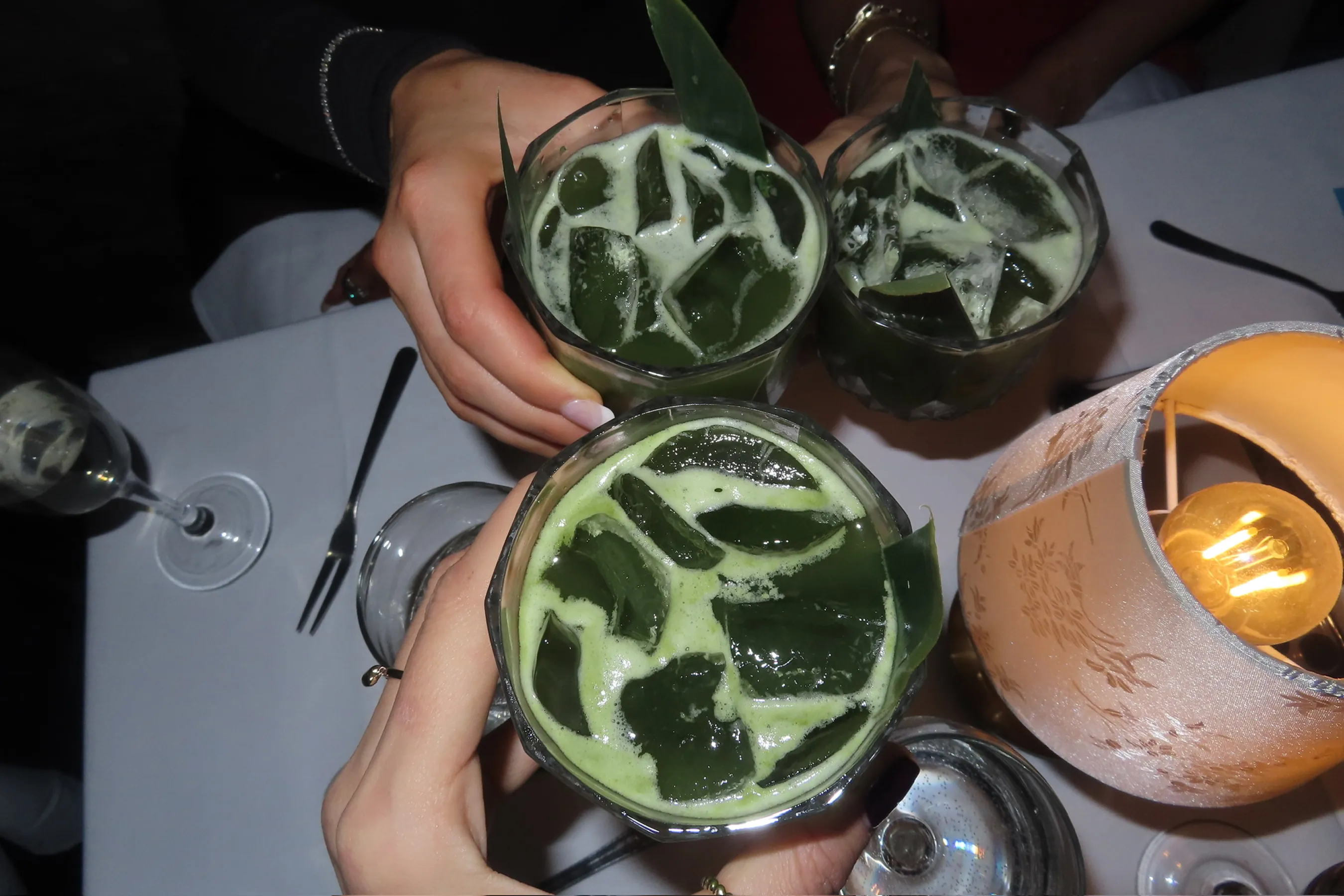 Close-up of three hands clinking faceted cocktail glasses filled with vibrant green drinks over ice, garnished with fresh leaves. A softly lit table lamp and elegant dinner setting in the background create a warm, celebratory atmosphere at an intimate evening event.