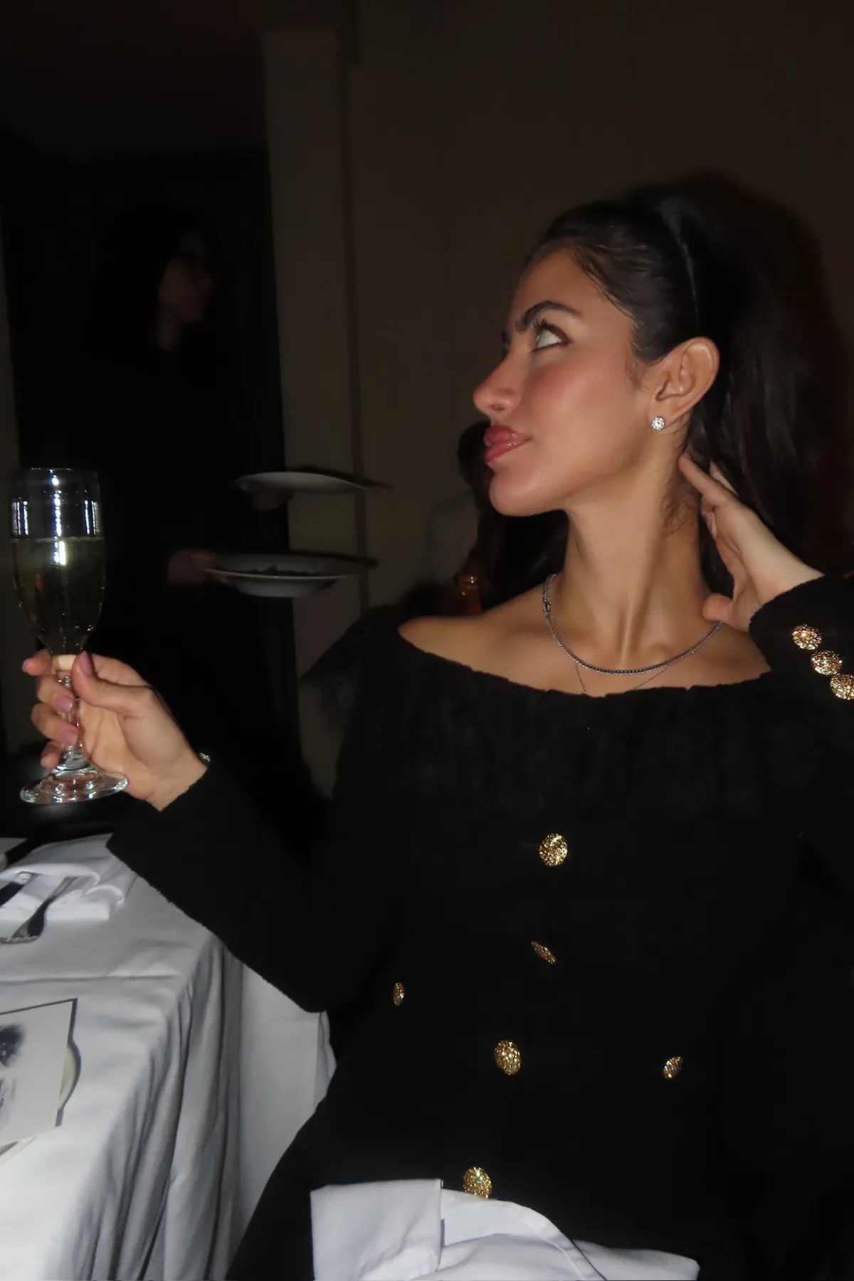 @therealsahd A woman seated at an elegant dinner table holds a champagne flute while looking to the side, wearing an off-the-shoulder black dress with gold button details. The softly lit restaurant setting and white table linens create a refined, intimate evening atmosphere.