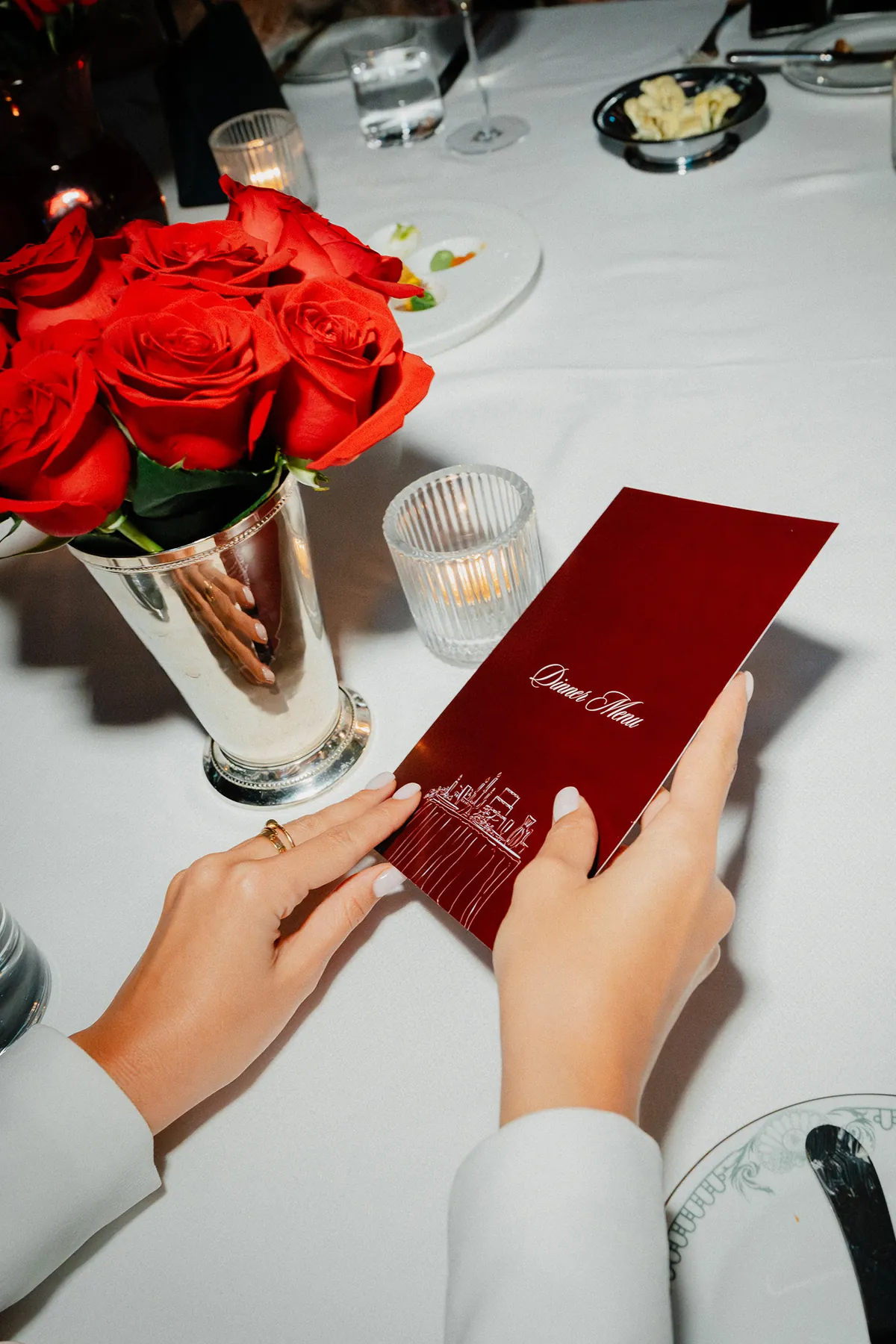 Storytime Creator holding a red dinner menu at a beautifully set table, decorated with a silver vase of red roses, candlelight, and polished dinnerware at Maison Barnes.