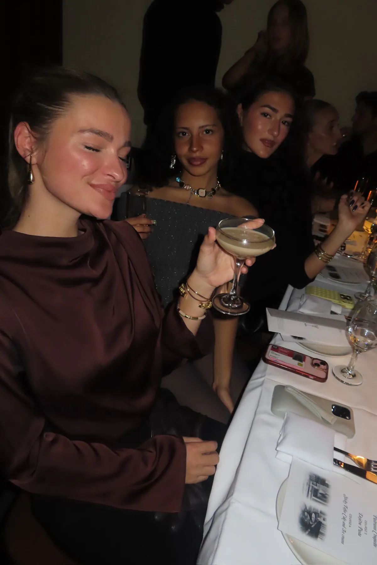 @carlabelleeee @ivycatrett and @corinnecll seated at an intimate dinner table, dressed in elegant evening attire and holding cocktails. Soft, low lighting and white table linens create a chic, upscale ambiance as guests enjoy drinks and conversation during the event.