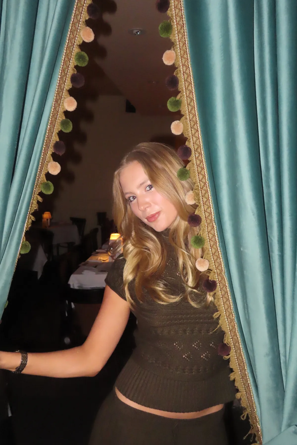 @mollymattporter parts teal velvet curtains trimmed with decorative pom-pom details, revealing a softly lit, elegant dining room behind her. She wears a fitted dark top and poses in the doorway, capturing the intimate and theatrical ambiance of the Storytime dinner at Déjà Vu.