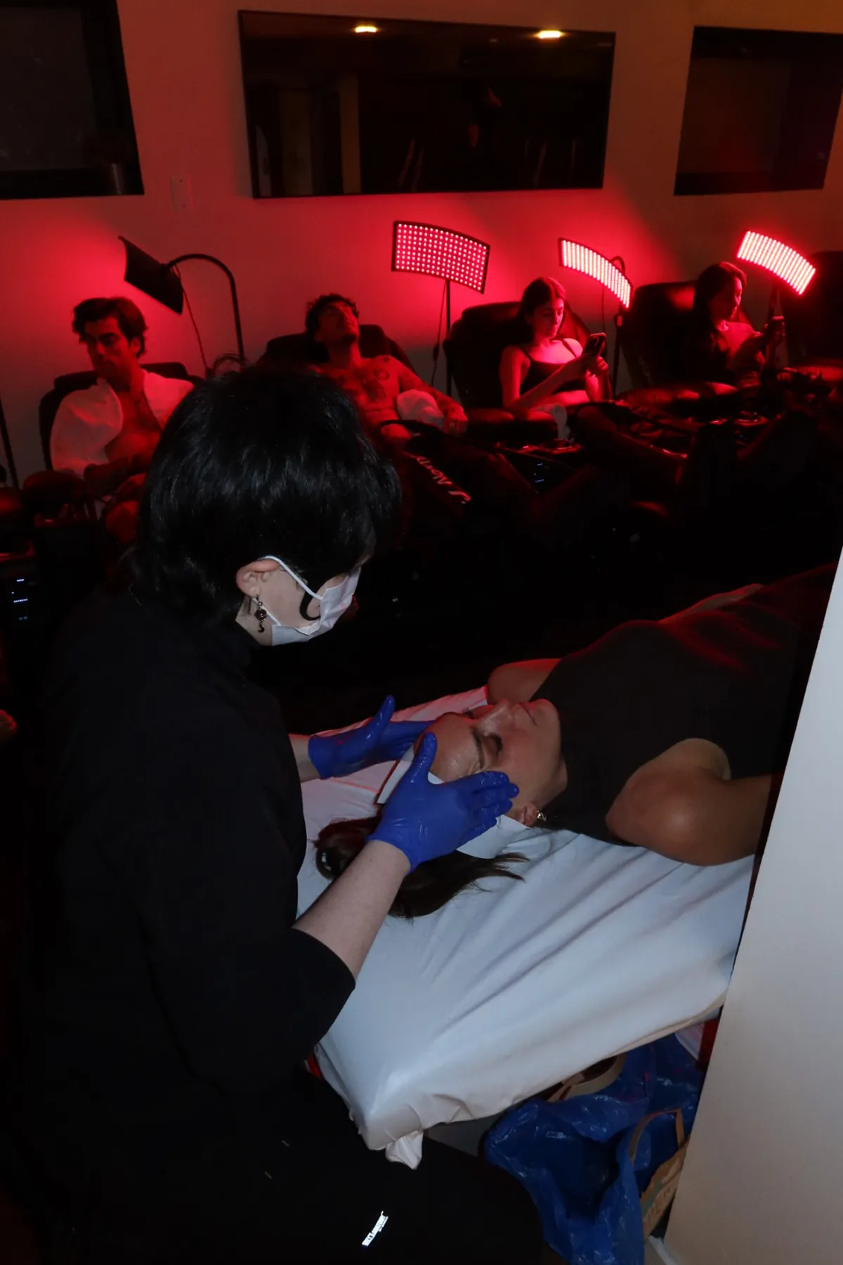 A guest receives a facial treatment by Glowbar at Kove Studio while others relax in reclining chairs under red light therapy panels. The dim, wellness-focused setting highlights recovery treatments and restorative experiences during the Storytime Wellness Reset Event.