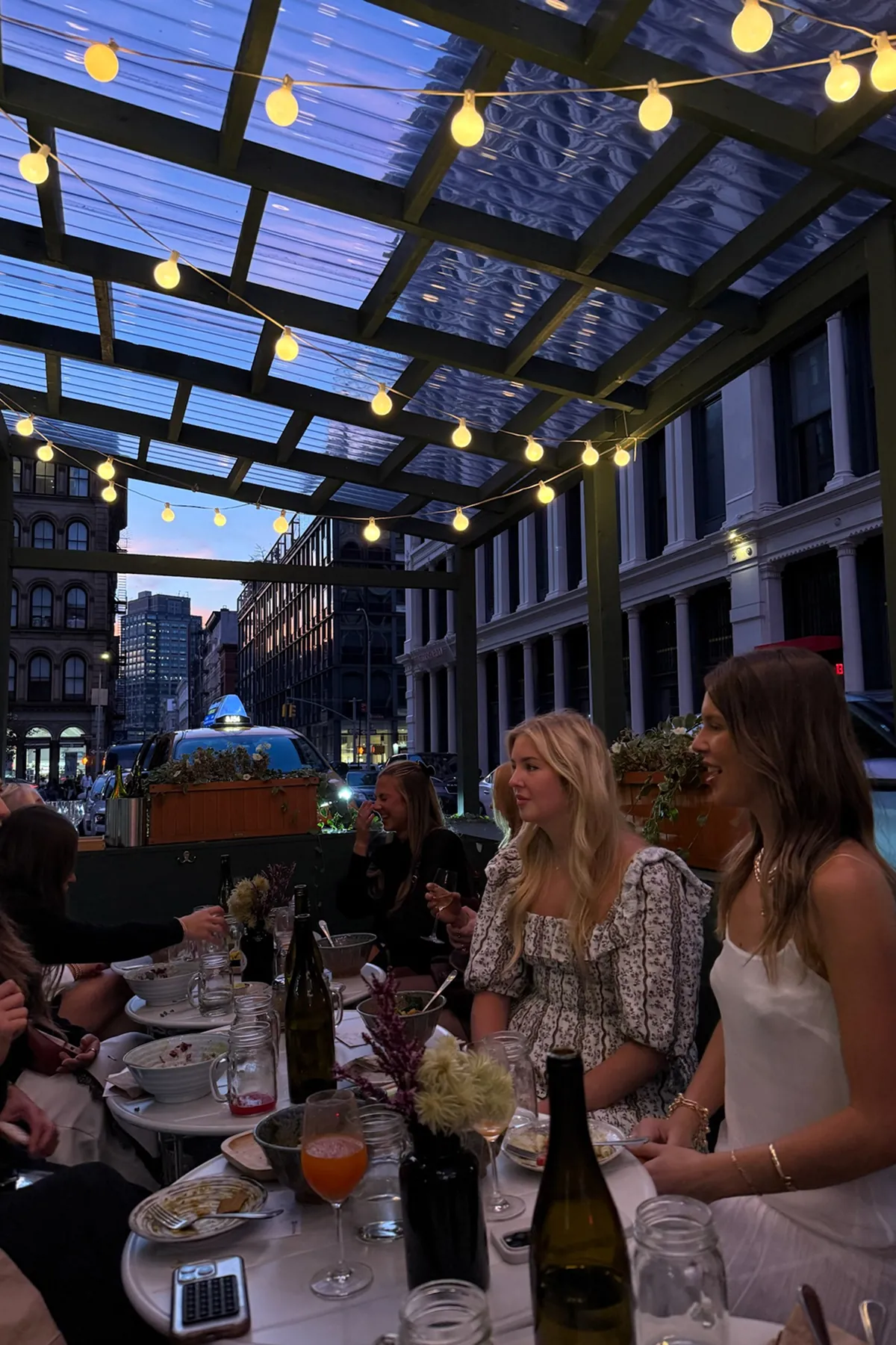 Storytime creators seated at a long outdoor dinner table at Le Botaniste under warm string lights, surrounded by dishes, drinks, and city buildings at dusk. The scene captures a cozy, intimate gathering focused on connection and conversation.