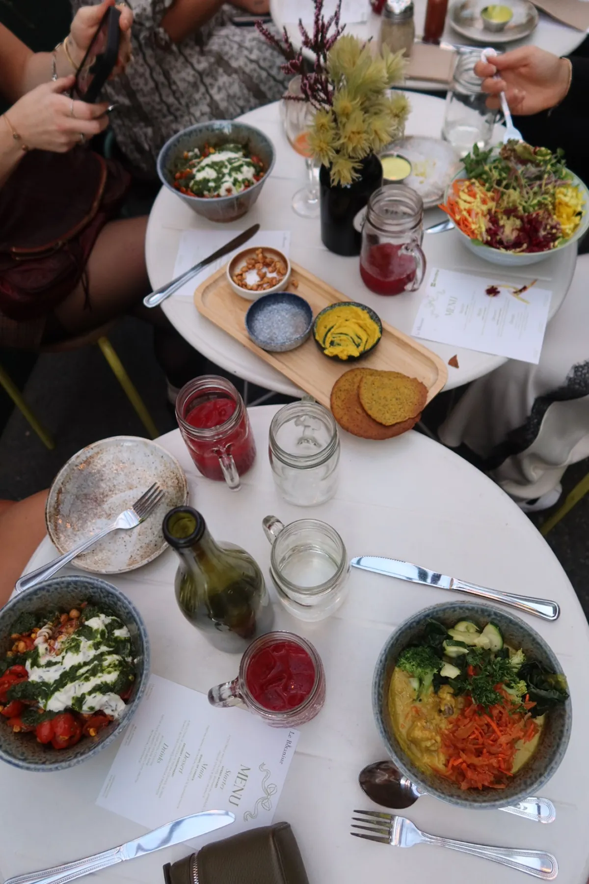 Overhead view of a Le Botaniste x Storytime dinner table filled with colorful plant-based bowls, hummus spreads, drinks in mason jars, fresh flowers, and guests capturing photos of their dishes.