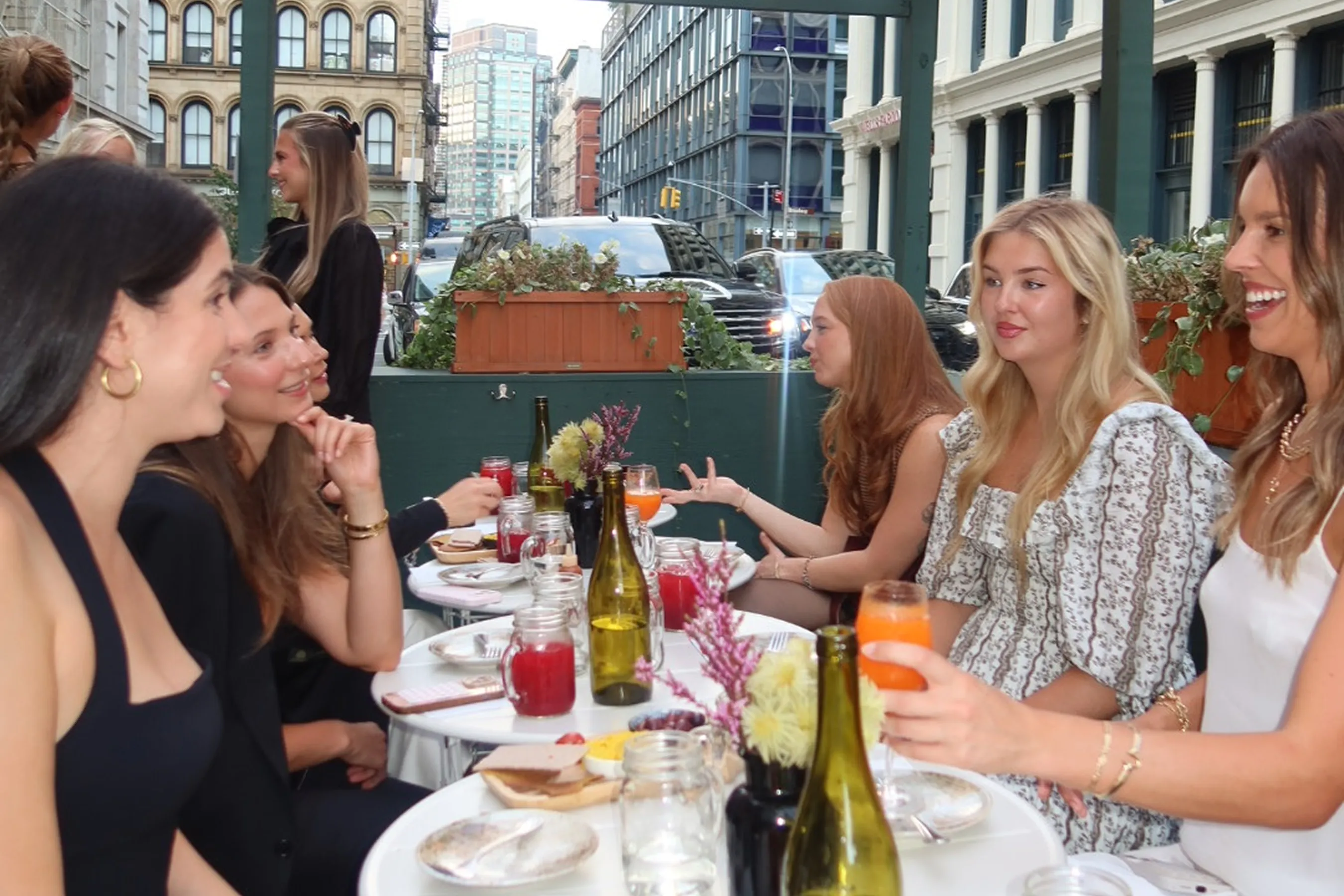 Storytime creators @allynissan, @carolinecorsello, @hannah.detwiler, @meribellekate, @ms.joynavarro, @elevatedwithelaine, @celestegoyena, @katie.overholt, @sydneyyhoover and @lilyystewart enjoying an outdoor dinner at Le Botaniste, seated at small round tables with plant-based dishes, botanical drinks, and flowers, while chatting against a lively SoHo street backdrop.