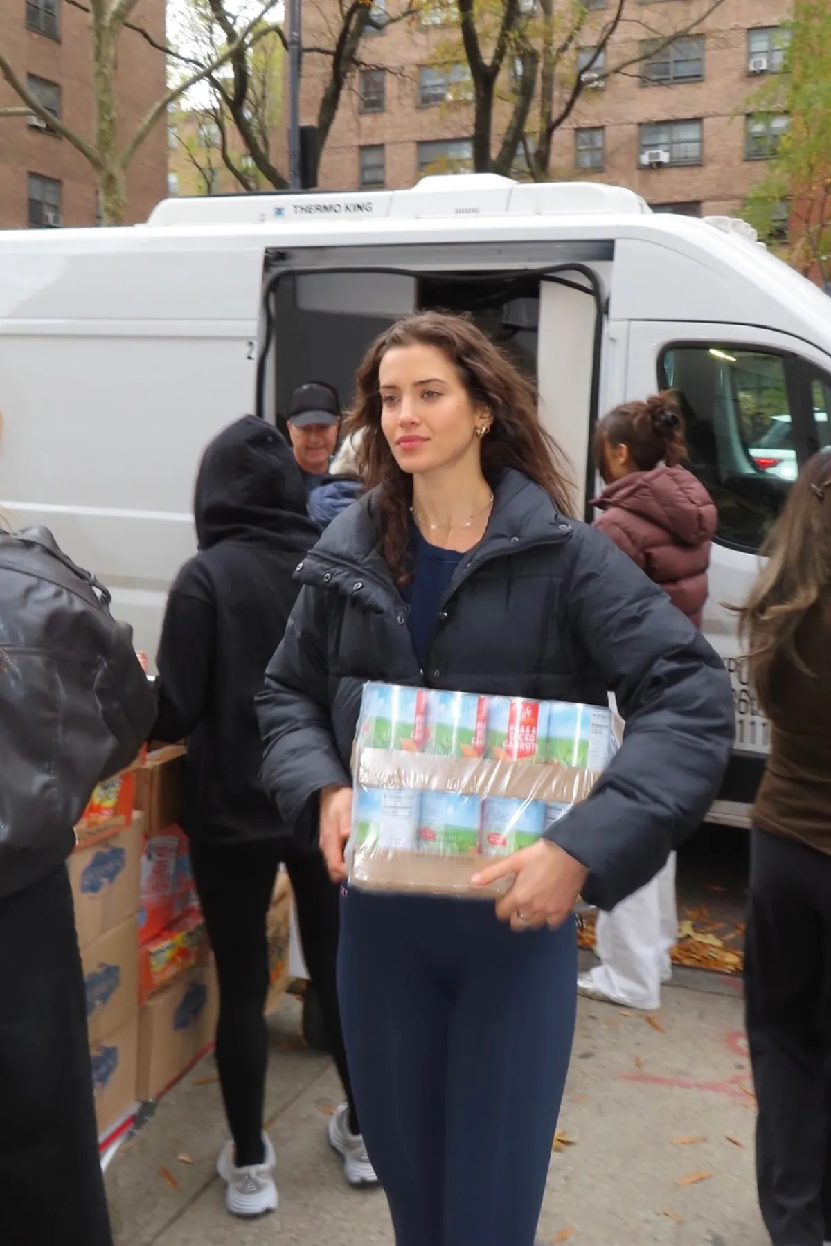 A Storytime creator carries a bundle of canned goods during a Thanksgiving volunteer event with The Migrant Kitchen Initiative, surrounded by other volunteers and a distribution van in the background.