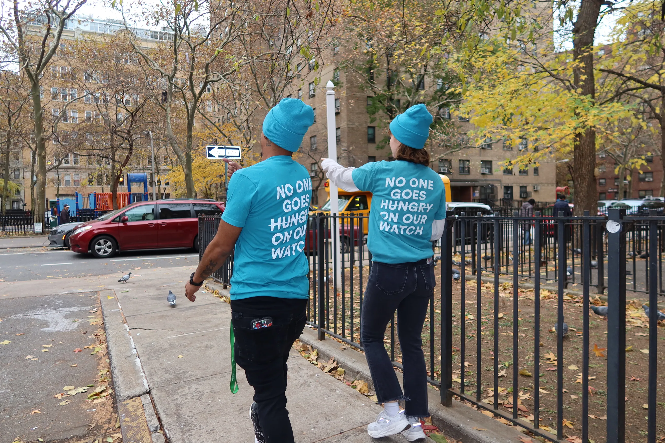 Two volunteers wearing matching turquoise shirts and beanies that read “No one goes hungry on our watch” walk through a New York neighborhood during the Thanksgiving distribution event with The Migrant Kitchen Initiative.