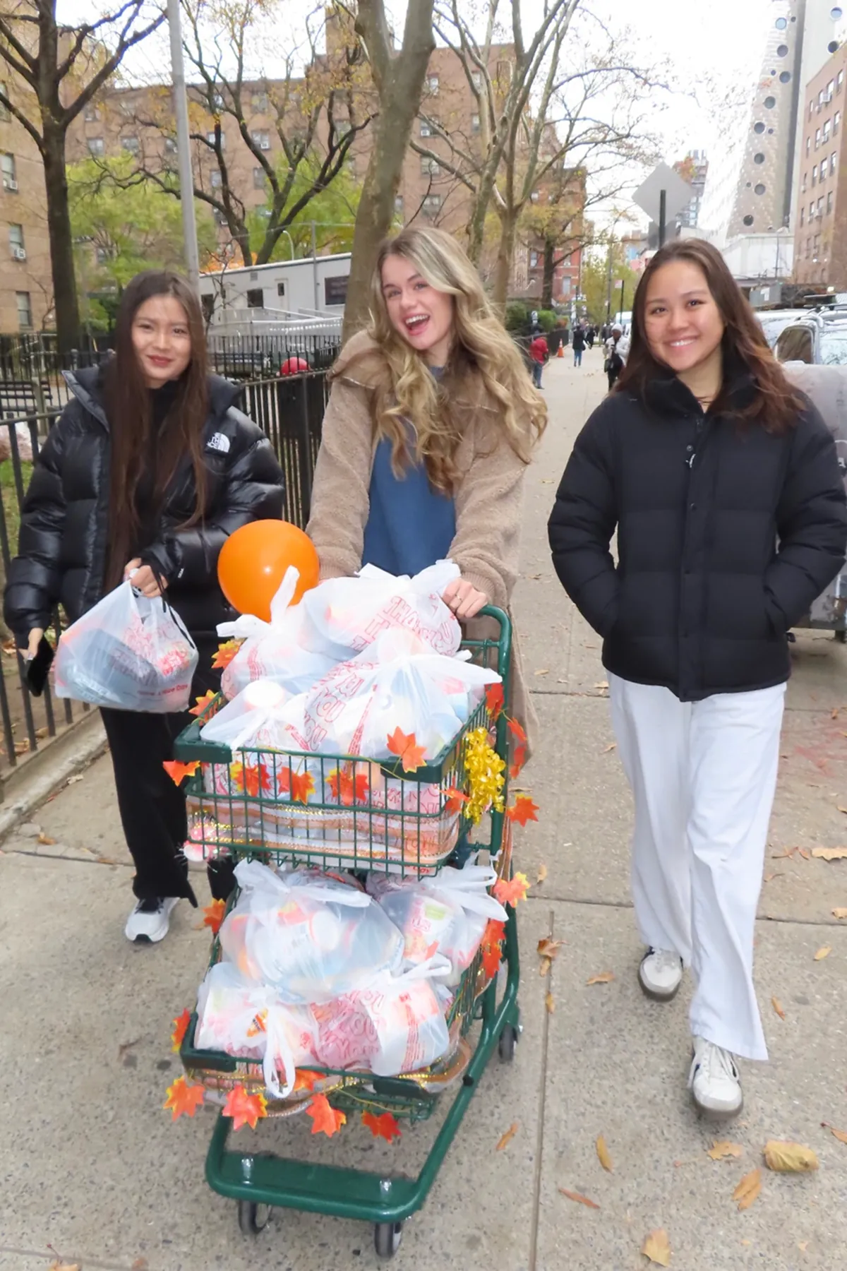 Three Storytime creators walk through a New York neighborhood while delivering Thanksgiving meal bags, pushing a decorated cart filled with food donations during The Migrant Kitchen Initiative volunteer event.