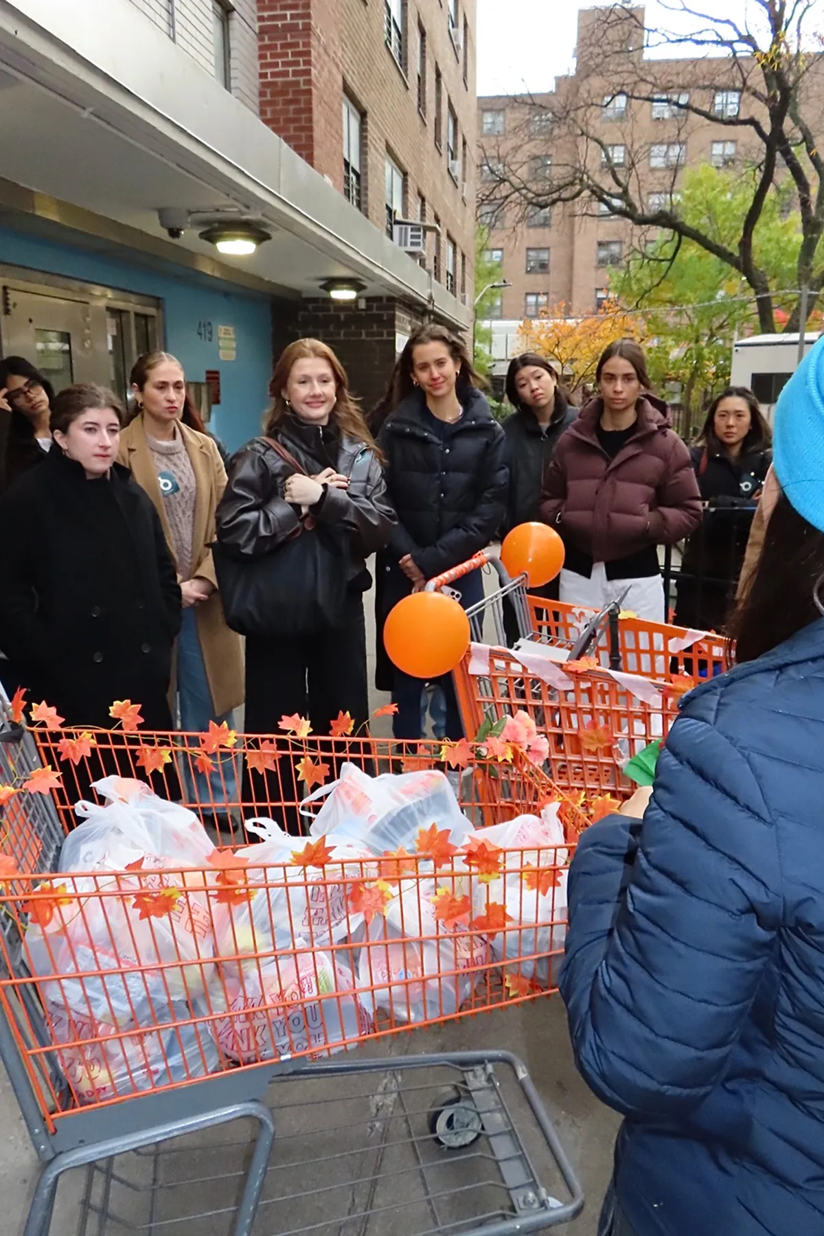 Storytime creators gather outdoors during a volunteer event with The Migrant Kitchen Initiative, listening attentively beside orange carts decorated with fall leaves and balloons and filled with Thanksgiving meal bags ready for distribution.