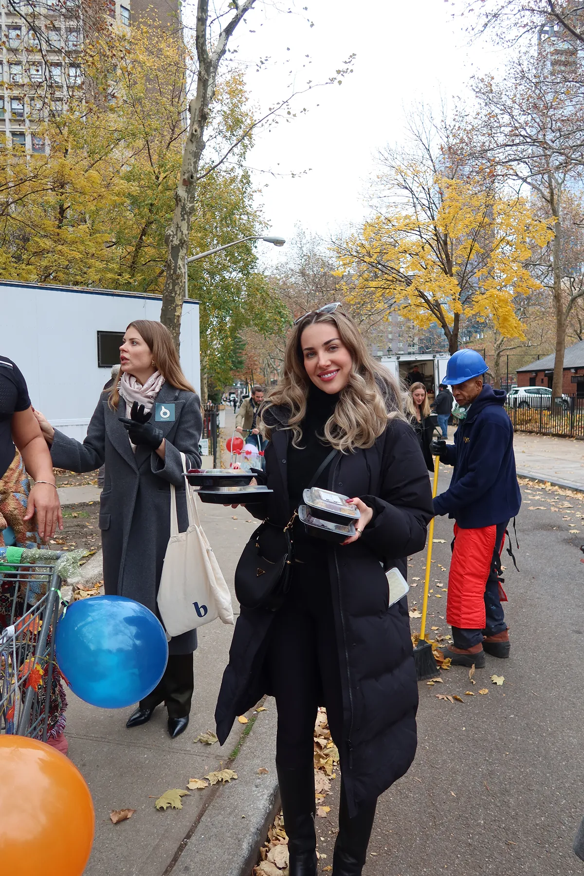 A Storytime creator smiles while carrying Thanksgiving meal trays during a volunteer event with The Migrant Kitchen Initiative, surrounded by fellow volunteers distributing food in a New York neighborhood.