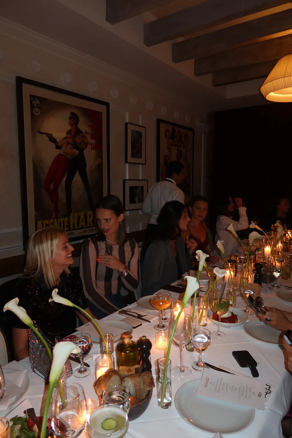 @josiejabs @anawolfermann @levilomey @gabeesco @catafreer @madsmaley @catafreer @elizabethfortmeyer @sally_carden @vito_9999 @ellie.forthegirls @isabellegraff @hannahwang @allinaai   @esraaguursoy @vikk.vas @avagparker @abbieisearley   @jennalitner @umiakiyoshi   @crumbsofnyc  @peachmangojuice   @nycbutglutenfree @mollymyattporter @miaachaloub   @lolanellie__   @madsmaley @josiejabs @zoeapple_ @uesthings @samnoyer @nycfoodfaves  @vito_9999     @taylordonoghuee @andrewpolo_ @treed_fl @theeuropeankid at a candlelit dinner table at Mamo's private dining room in New York, decorated with white calla lilies, bread, and wine glasses. Guests dressed in chic evening outfits sit closely together, talking, laughing, and enjoying cocktails in a warm, intimate dining room with vintage movie posters on the walls.