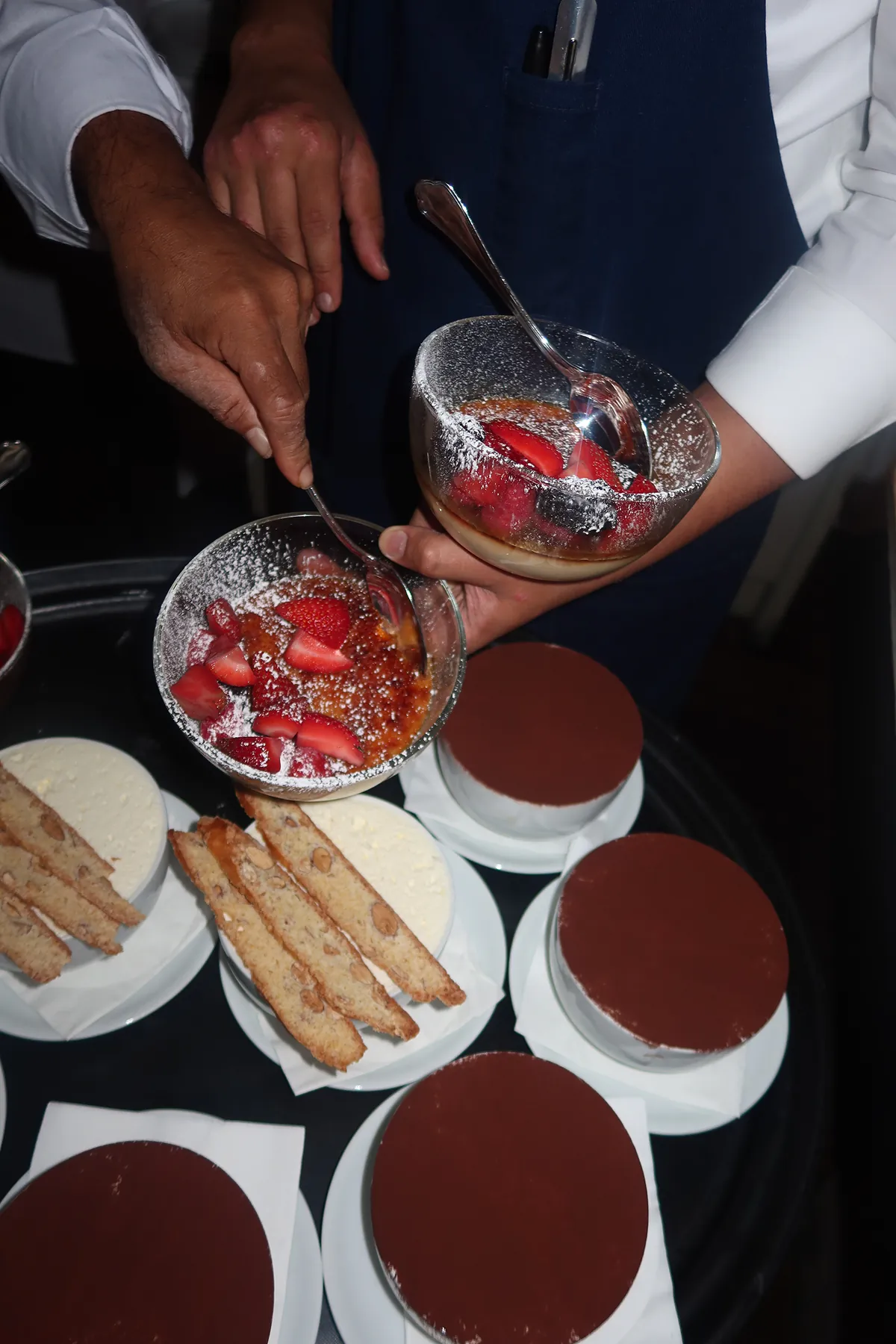 Waiters at Mamo serving crème brûlée topped with fresh strawberries, surrounded by plated desserts and biscotti. 