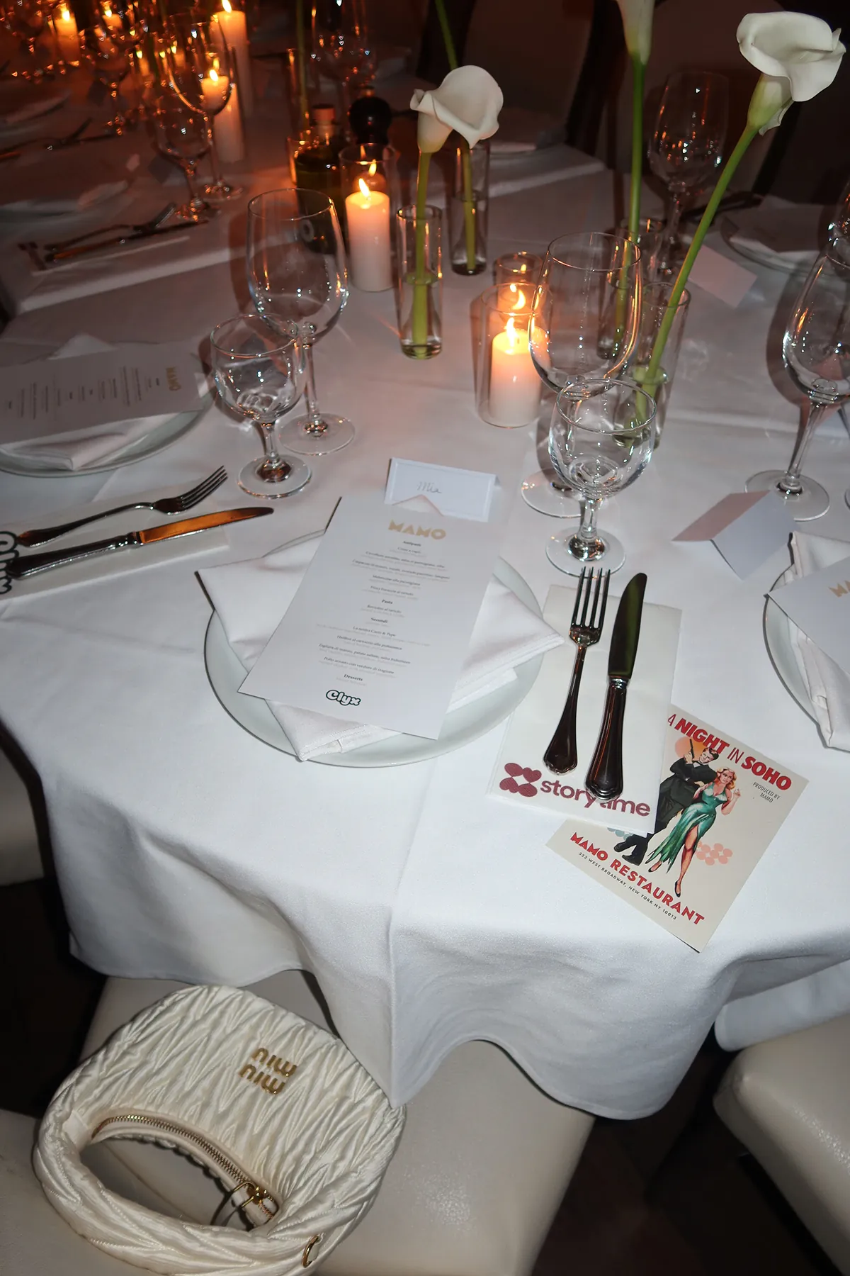 Elegant dinner table setup at Mamo restaurant with white linens, candles, calla lilies, multiple wine glasses, a printed menu, and a Storytime event card, with a white Miu Miu bag placed on the chair.