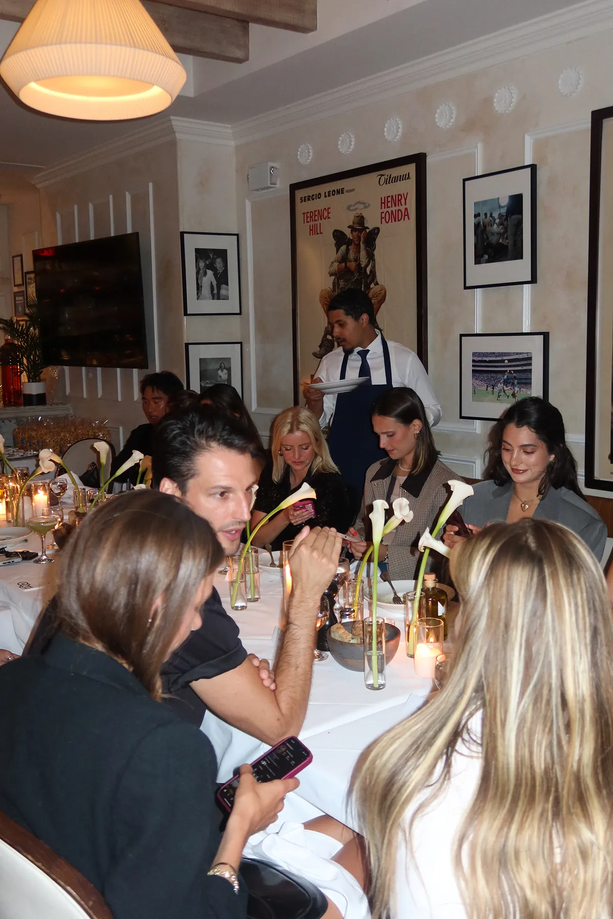 @josiejabs @anawolfermann @levilomey @gabeesco @catafreer @madsmaley @catafreer @elizabethfortmeyer @sally_carden @ellie.forthegirls @isabellegraff @hannahwang @allinaai   @esraaguursoy @vikk.vas @avagparker @abbieisearley @jennalitner @crumbsofnyc @nycbutglutenfree @mollymyattporter @miaachaloub @lolanellie__ @madsmaley @josiejabs @uesthings @samnoyer @nycfoodfaves @taylordonoghuee @andrewpolo_ @treed_flseated @levilomey at a long candlelit dinner table at Mamo restaurant during a Storytime event, with calla lily centerpieces, framed artwork on the walls, and a server carrying plates.