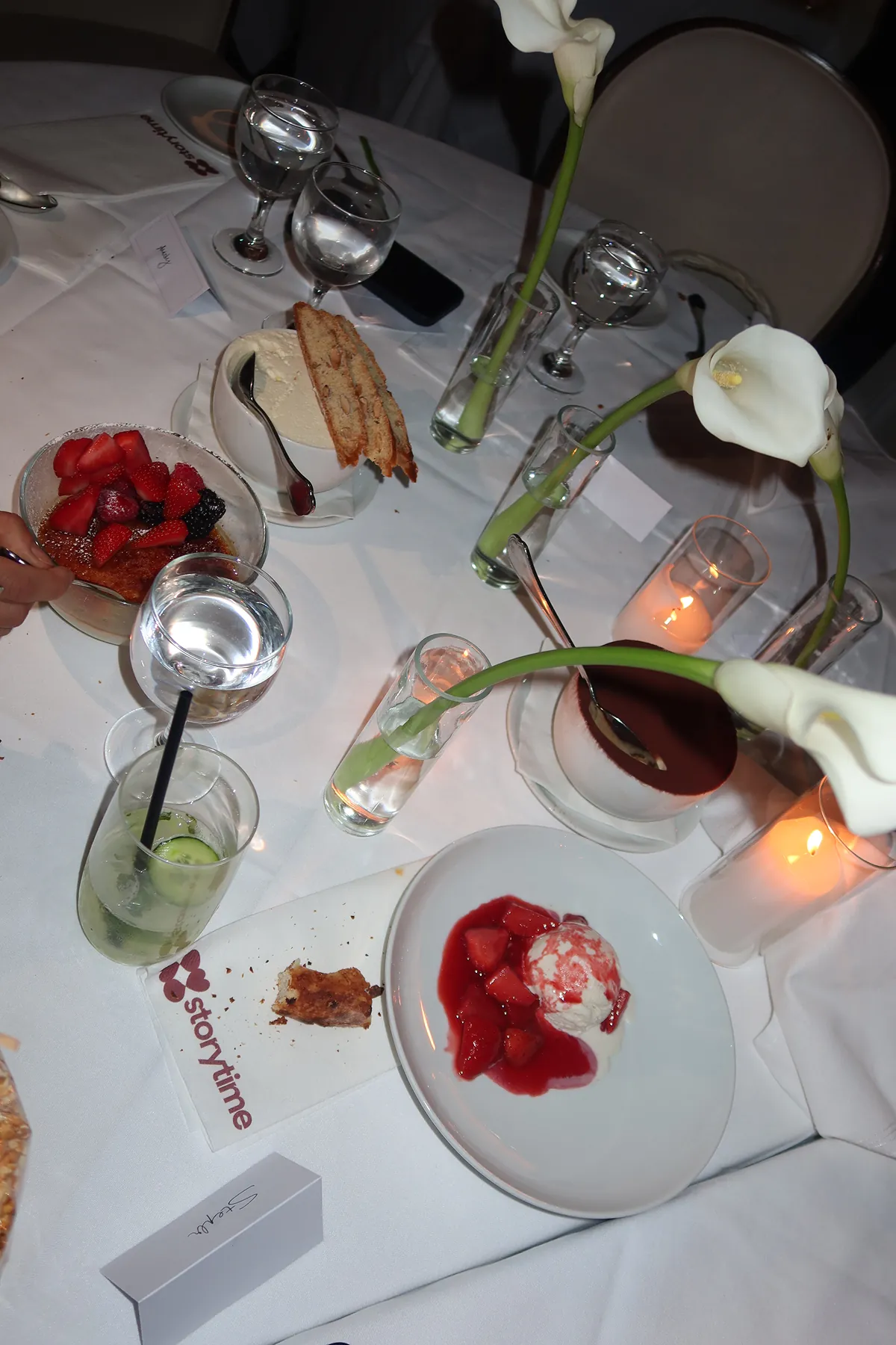 abletop at a Mamo x Storytime dinner featuring assorted desserts including crème brûlée with berries, tiramisu, and strawberry-topped ice cream, surrounded by candles, calla lily centerpieces, and drinks on a white tablecloth.