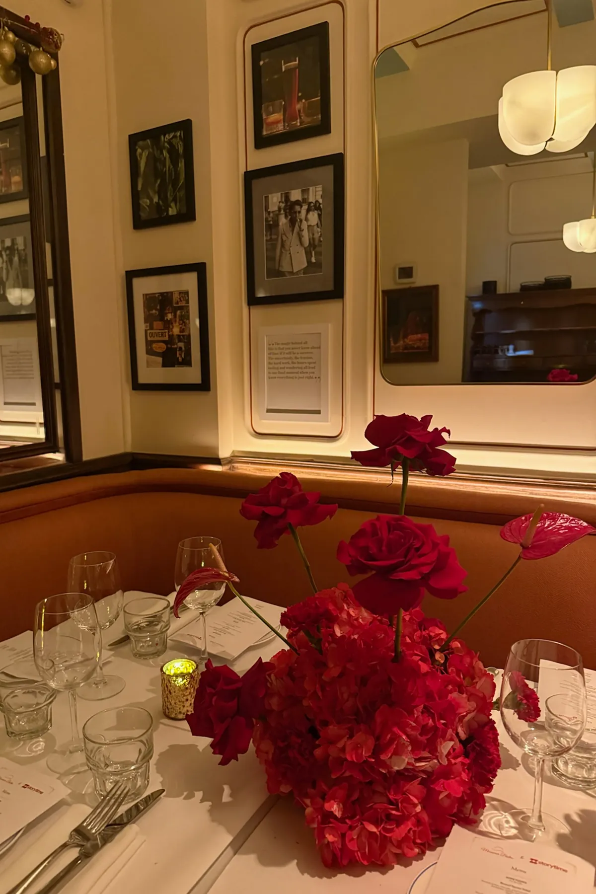 An intimate holiday table setting at Monsieur Bistro featuring vibrant red floral centerpieces from @piroflowers by @natalyapiro , glowing candlelight, wine glasses, and elegant place settings. Framed artwork and warm lighting create a cozy, festive ambiance for the Storytime Christmas dinner event.