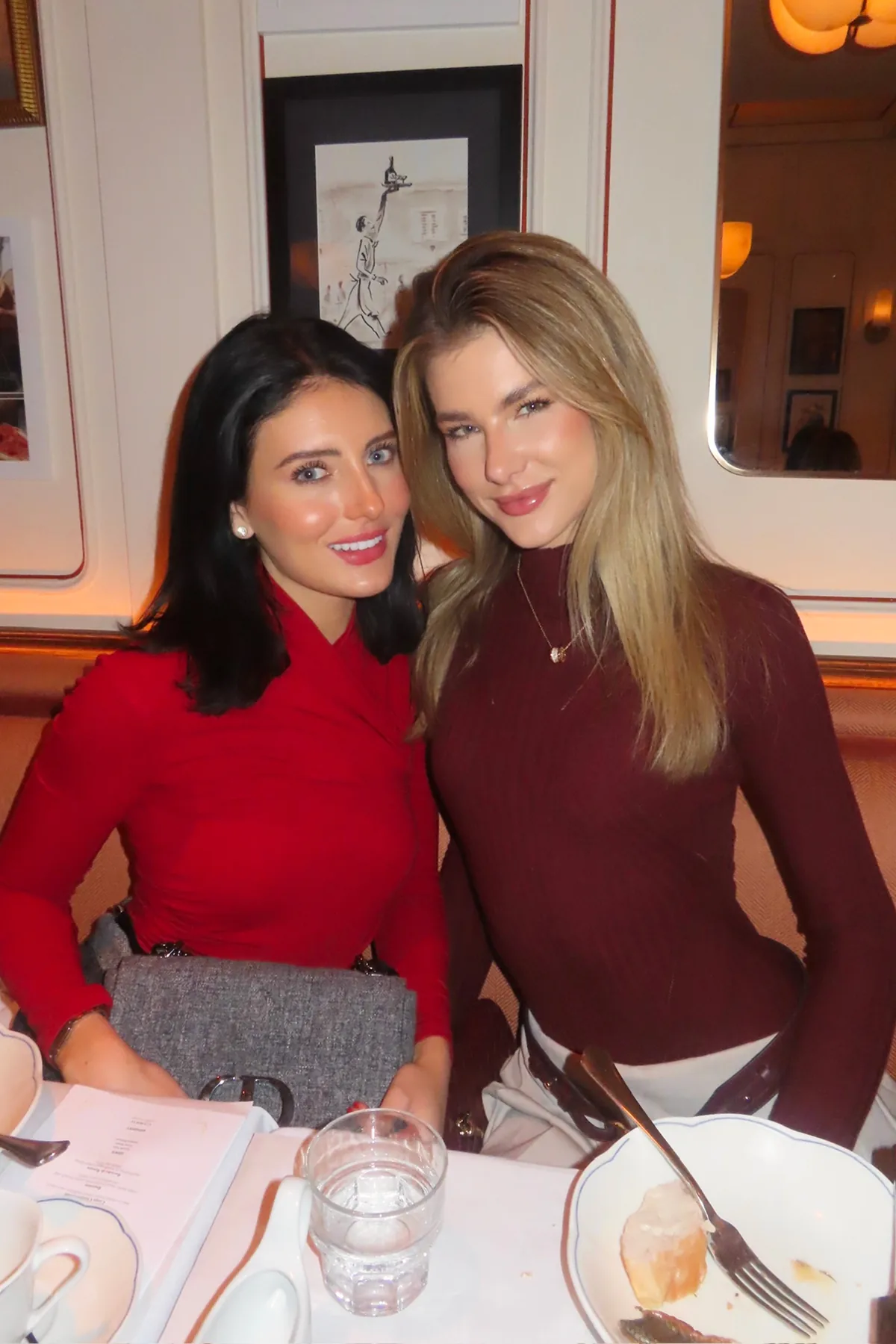 @jojo.n.chann and @jennadykstraa dressed in elegant red and burgundy attire pose closely together at a holiday dinner table inside Monsieur Bistro. Warm lighting, framed artwork, and refined place settings create a cozy, festive atmosphere during the Storytime Christmas dinner event.