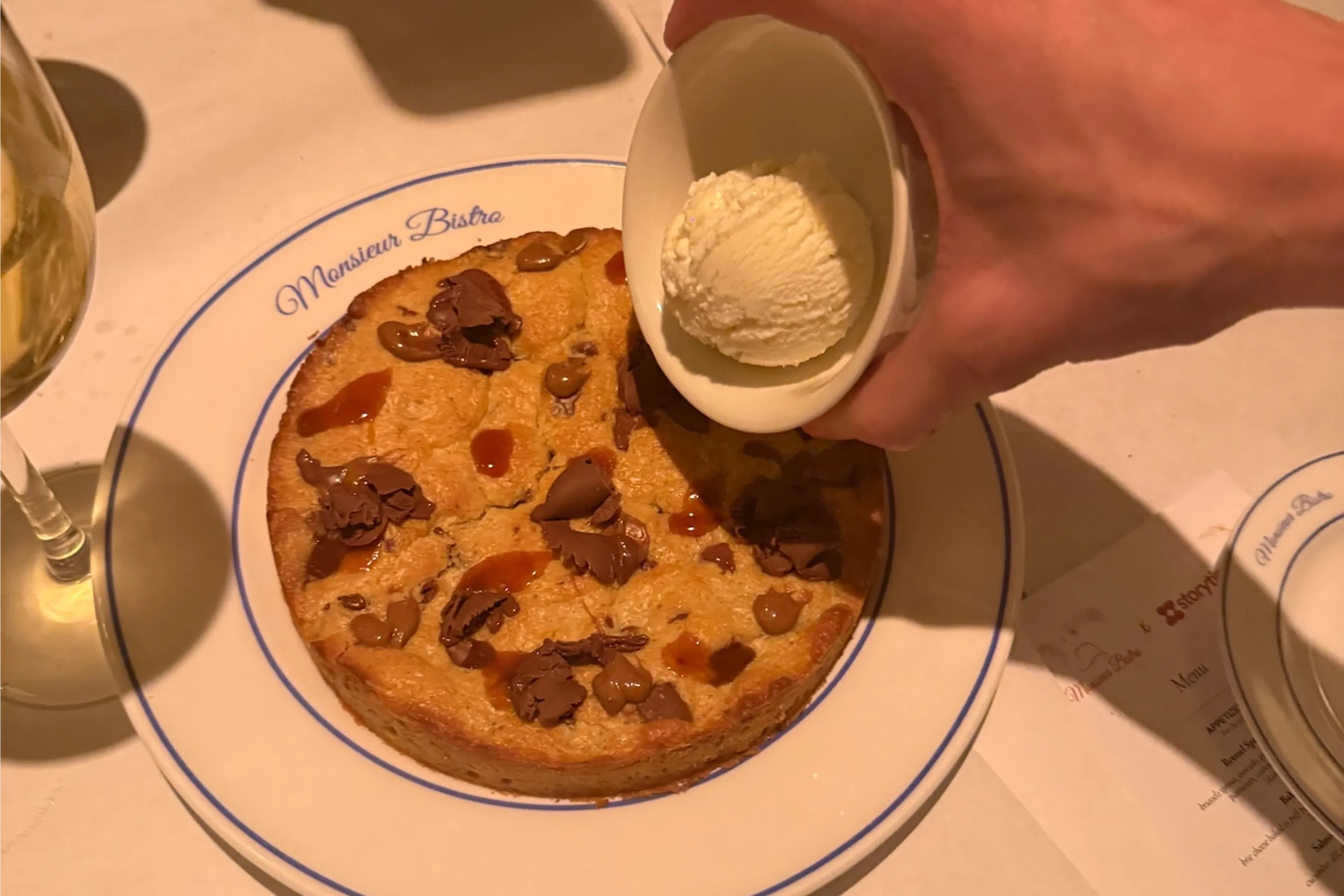 Close-up of the viral @monsieurbistro cookie dessert topped with chocolate shavings and caramel drizzle, served on a Monsieur Bistro plate. A scoop of vanilla ice cream is being added, highlighting the indulgent, festive dining experience at the Storytime Christmas dinner.