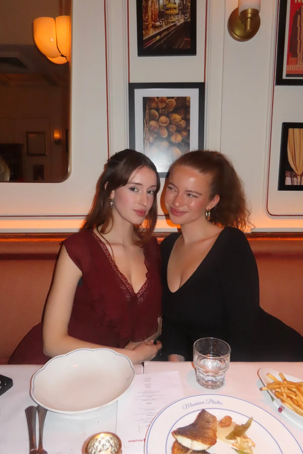 @aashleysuarez seated side by side on a leather banquette at Monsieur Bistro, dressed in elegant burgundy and black outfits. Warm lighting, framed artwork, and refined table settings create a cozy, festive atmosphere during the Storytime Christmas dinner event.