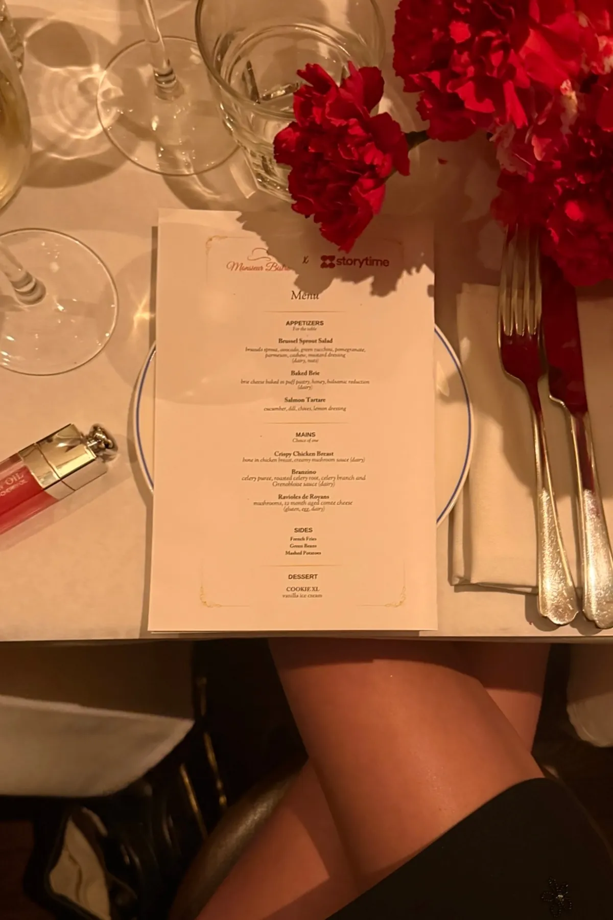 @nycbutglutenfree Overhead view of a curated Monsieur Bistro x Storytime menu placed on an elegantly set table with wine glasses, polished silverware, and vibrant red floral accents. Warm lighting highlights the refined holiday dining experience at the Storytime Christmas dinner event.