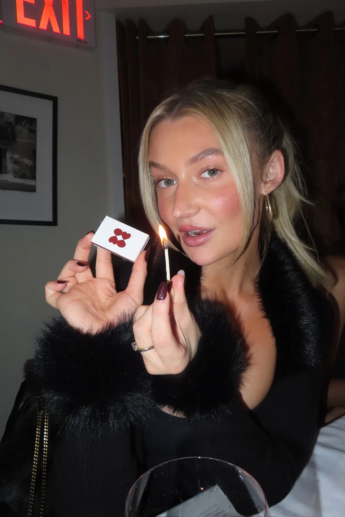 @itslaurenraymond holds a lit match in one hand and a Storytime-branded matchbox in the other. She is dressed in a black outfit with faux-fur sleeves and is seated at a dinner table, illuminated by warm indoor lighting.