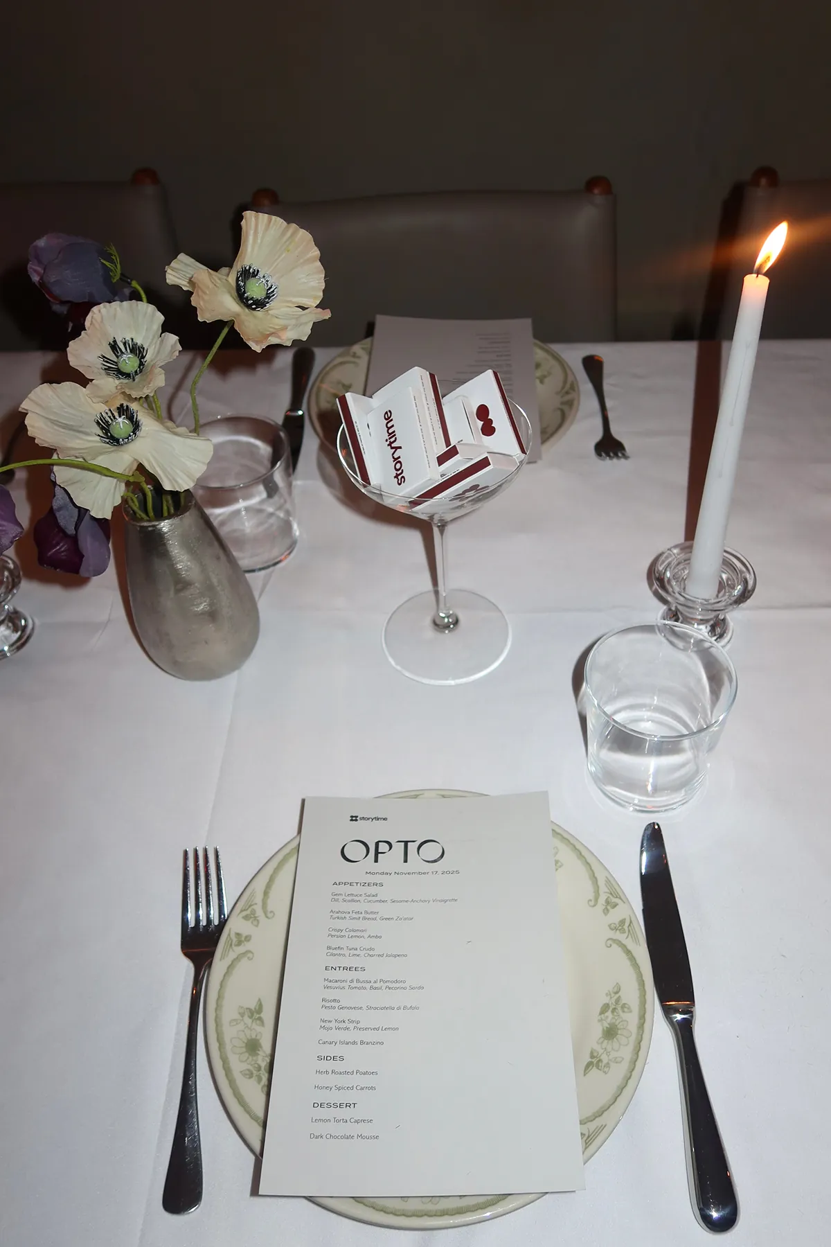 A candlelit dinner table at Opto featuring a Storytime-branded menu, vintage-style plates, a martini glass filled with Storytime matchboxes, and a vase of cream and purple flowers set from @apartement.387 for the Friendsgiving event.