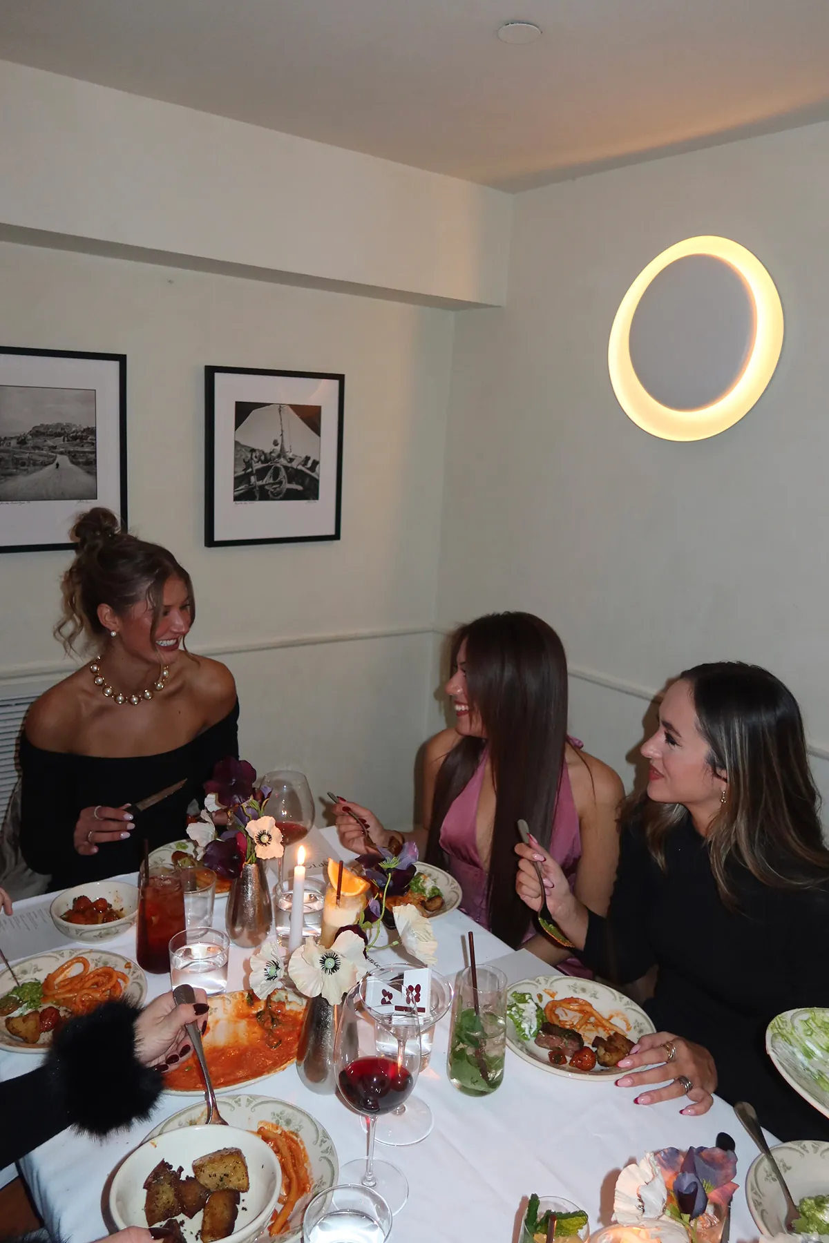@katie.overholt, @_taniagrover and @evitascoccia enjoying dinner at Opto in New York, sitting around a candlelit table with plates of Italian dishes, cocktails, and floral centerpieces during the Storytime Friendsgiving event.