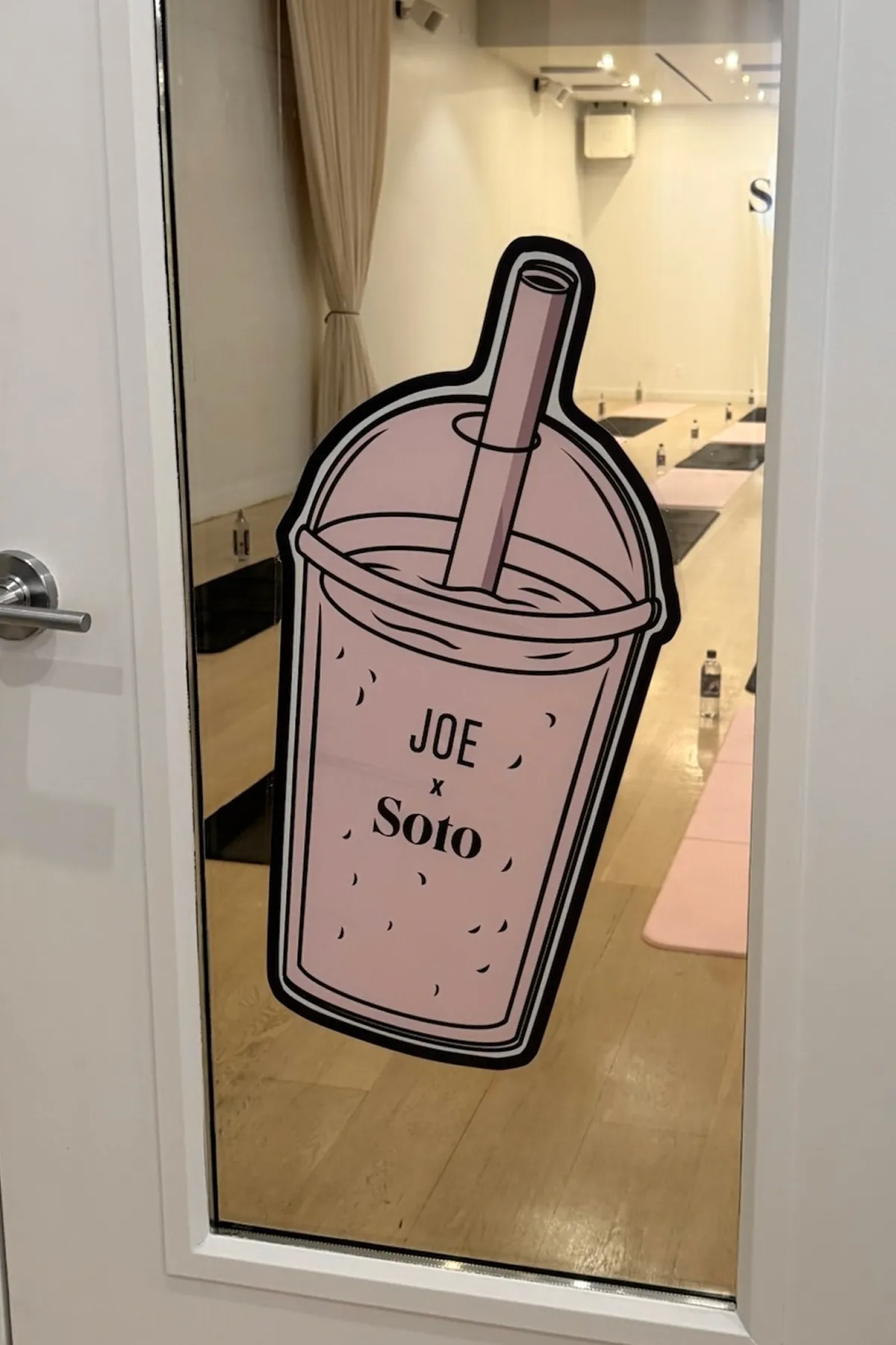 Large pink Soto Method in collaboration with Joe and The Juice sticker of a smoothie cup labeled “JOE x Soto” overlooking Soto Method fitness studio set up with pink and black mats and water bottles arranged for a class.