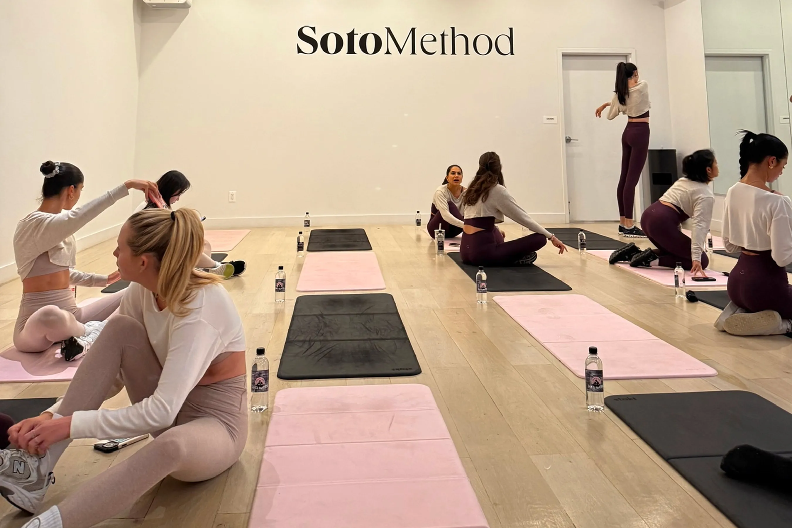 @alexis_loomans, @kate___marko, @andreaventurrr, @avb.fit, @emmawilliams18, @erika.dwyer, and @scopelcami stretching on pink and black mats at the Soto Method studio, preparing for a mat pilates class.