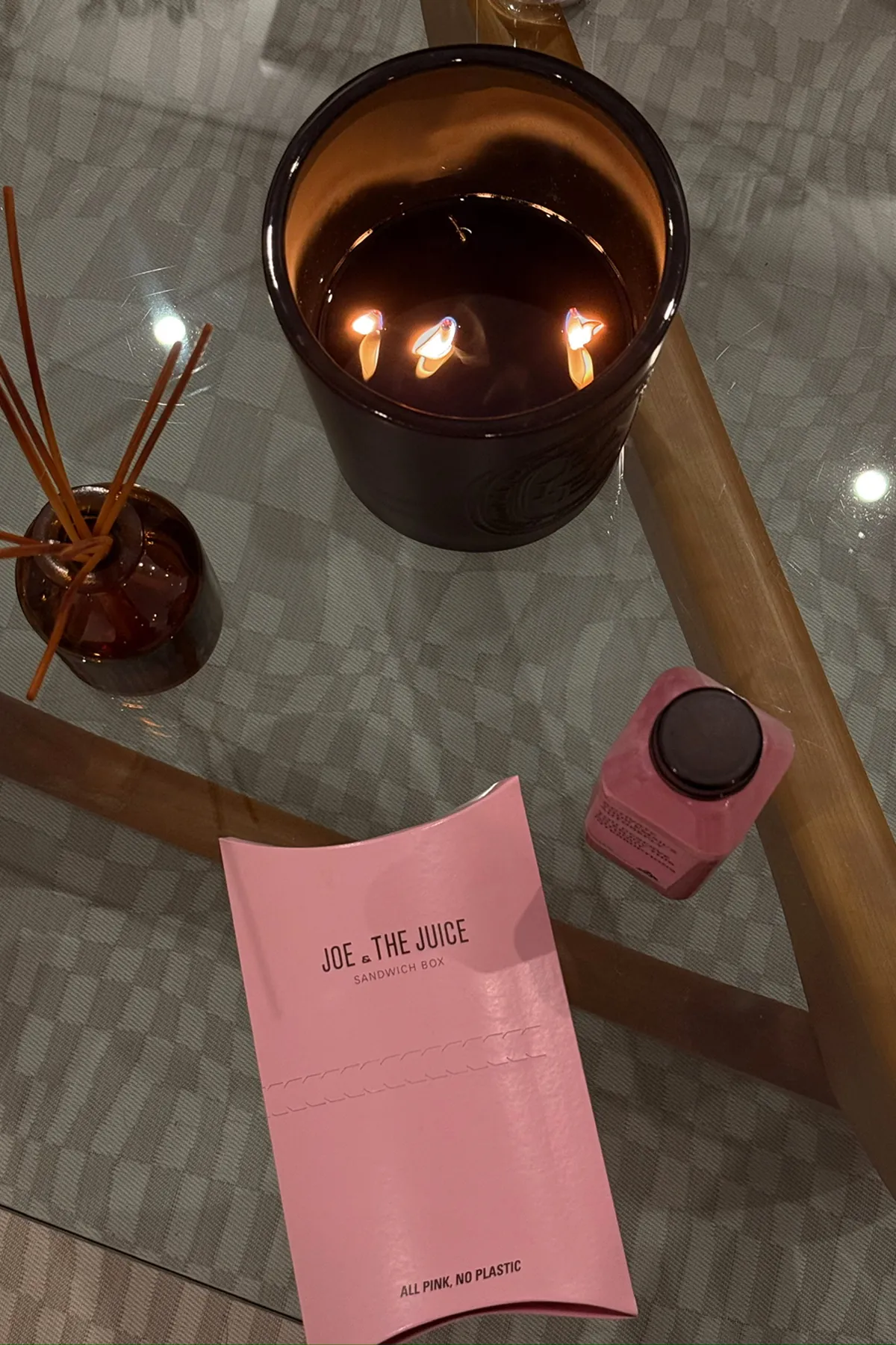 Pink Joe & The Juice sandwich box and a matching pink bottled juice placed on a glass table beside a burning three-wick candle and a small reed diffuser.