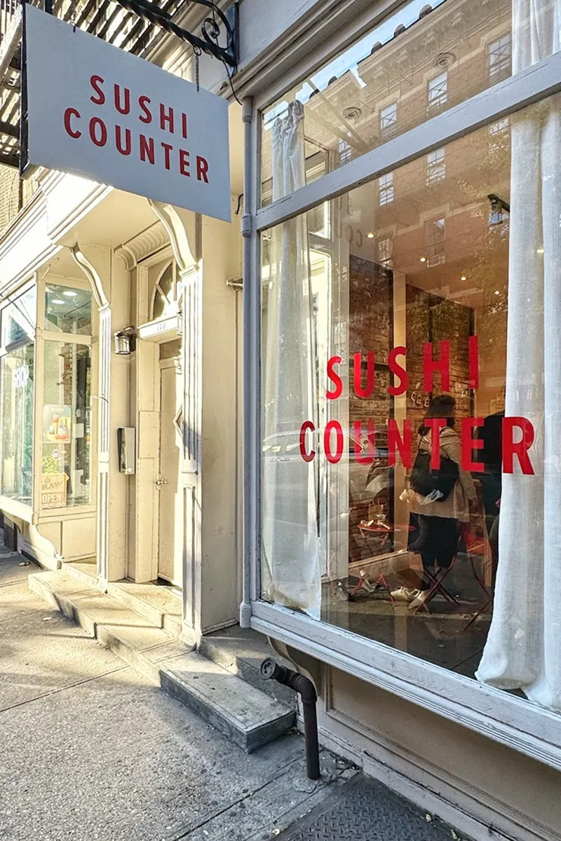 Street view of Sushi Counter’s storefront with large red logo on the window and hanging sign, showing the bright, minimalist exterior and reflections of the neighborhood.