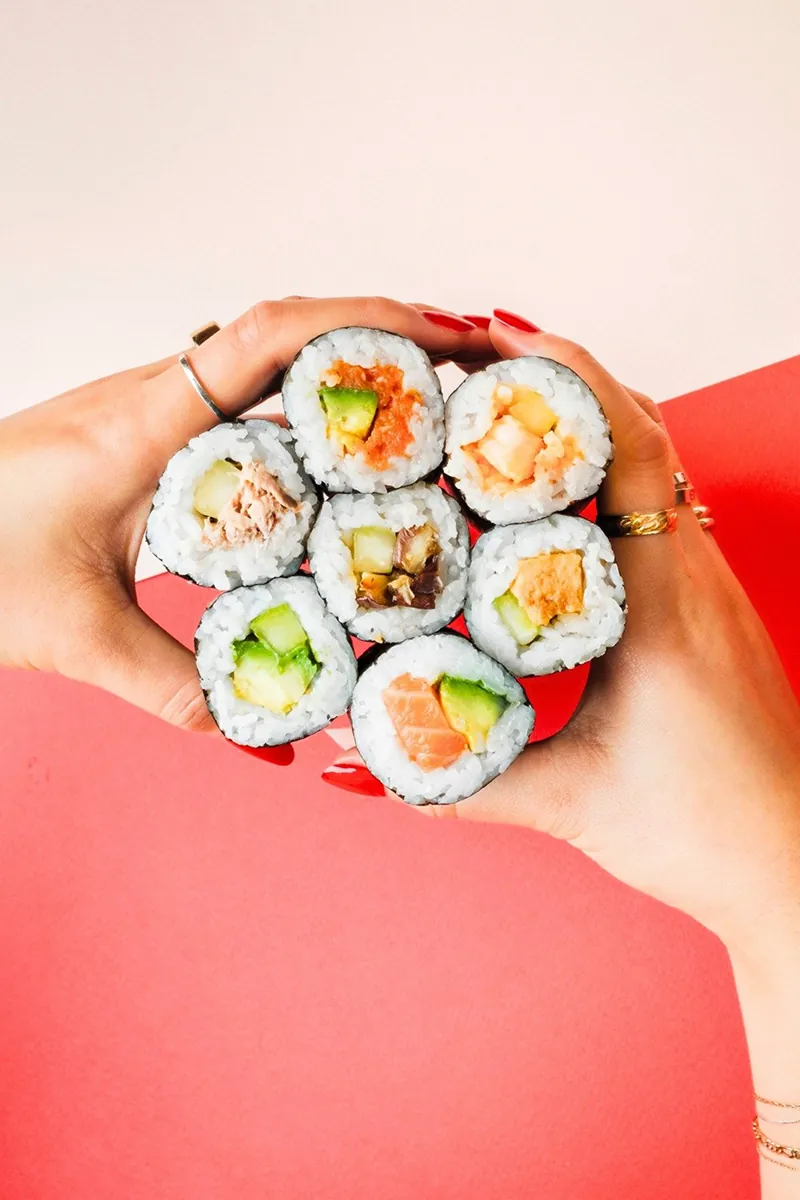 Close-up of hands holding a bundle of Australian sushi rolls from Sushi Counter NYC with fresh fillings like salmon, tuna, avocado, and cucumber against a pink and red background.