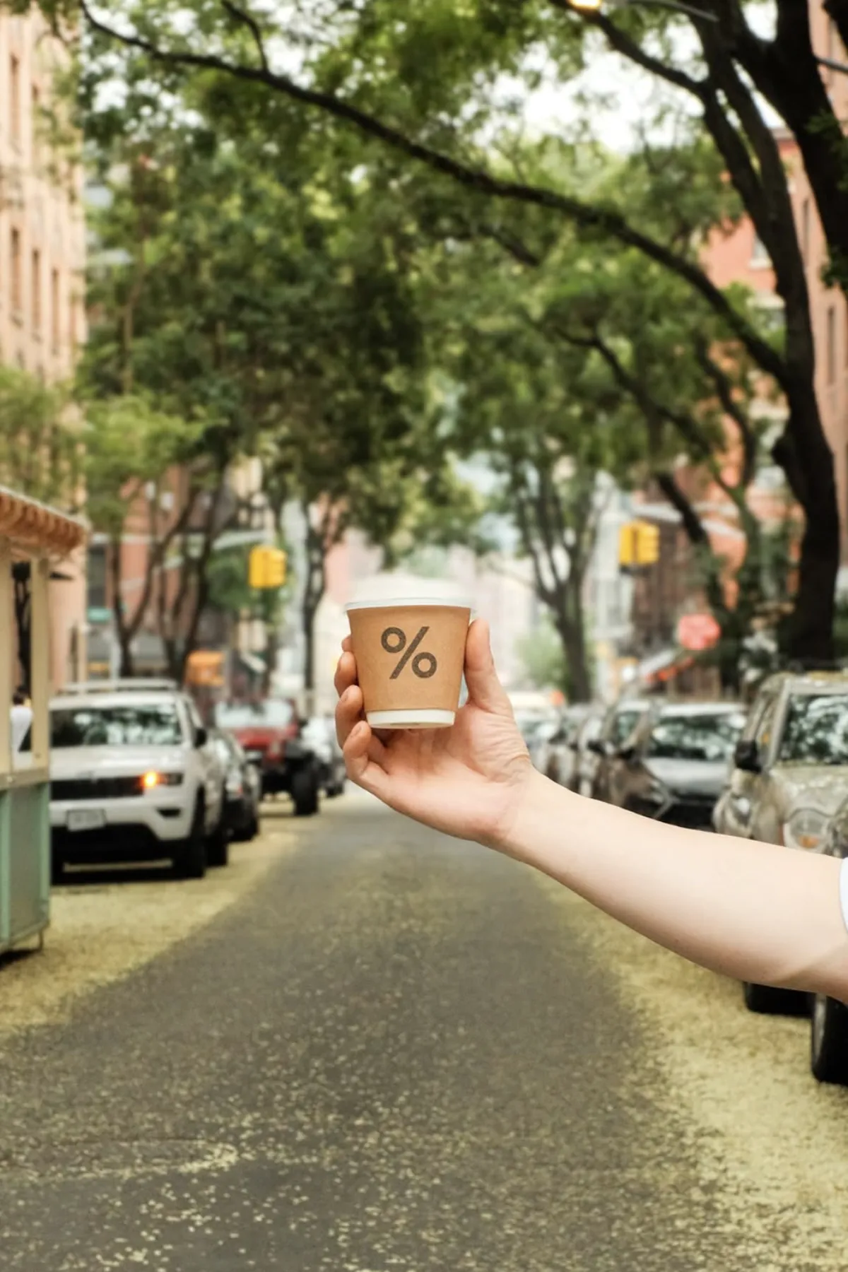 A hand holds a small % Arabica coffee cup in the middle of a tree-lined New York City street. Parked cars, soft natural lighting, and leafy branches frame the scene, highlighting the minimalist design and urban lifestyle aesthetic associated with the % Arabica café brand.